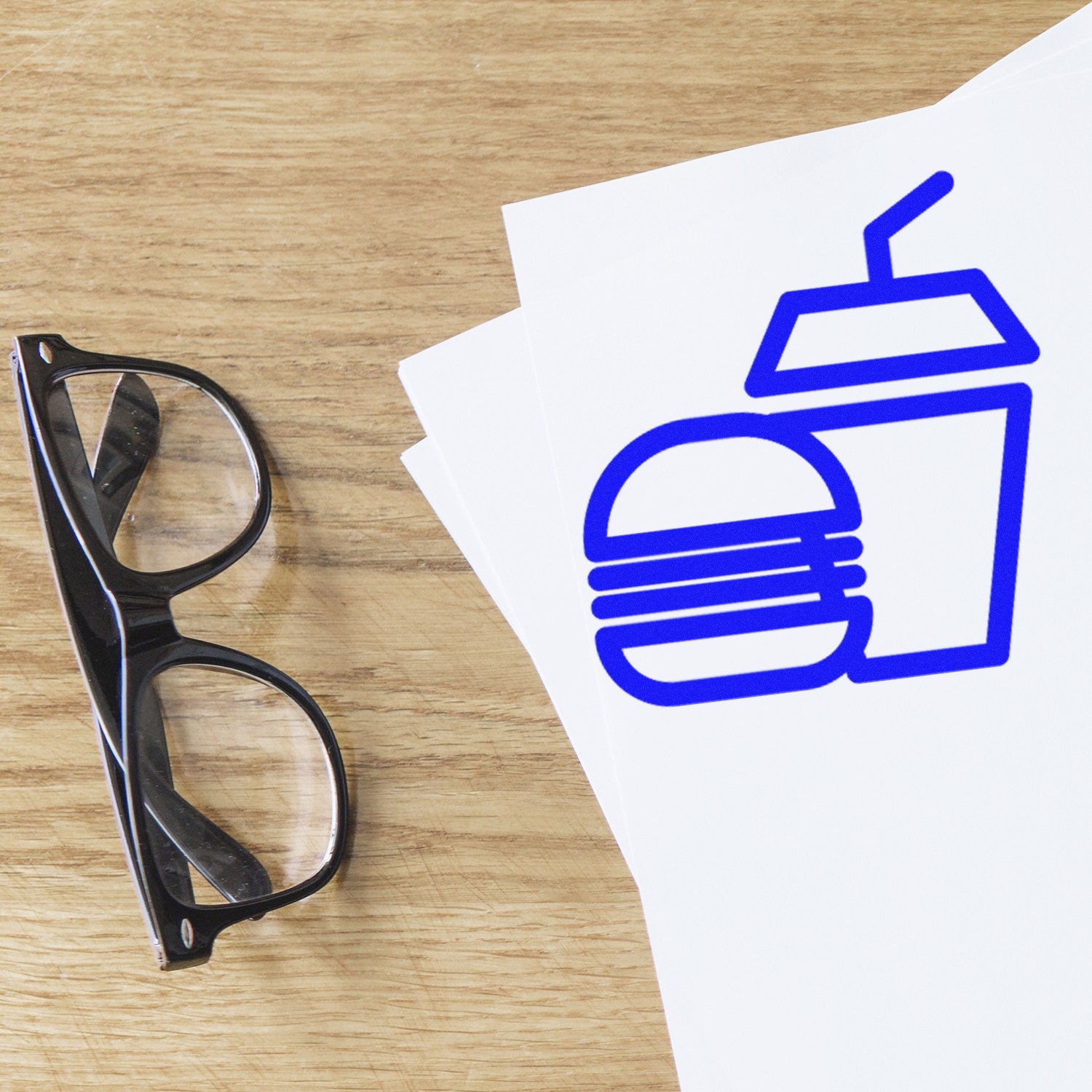 Wood Handle Burger Bliss Loyalty Punch Card Rubber Stamp on a wooden surface, next to black glasses and stamped paper with a blue burger and drink icon.