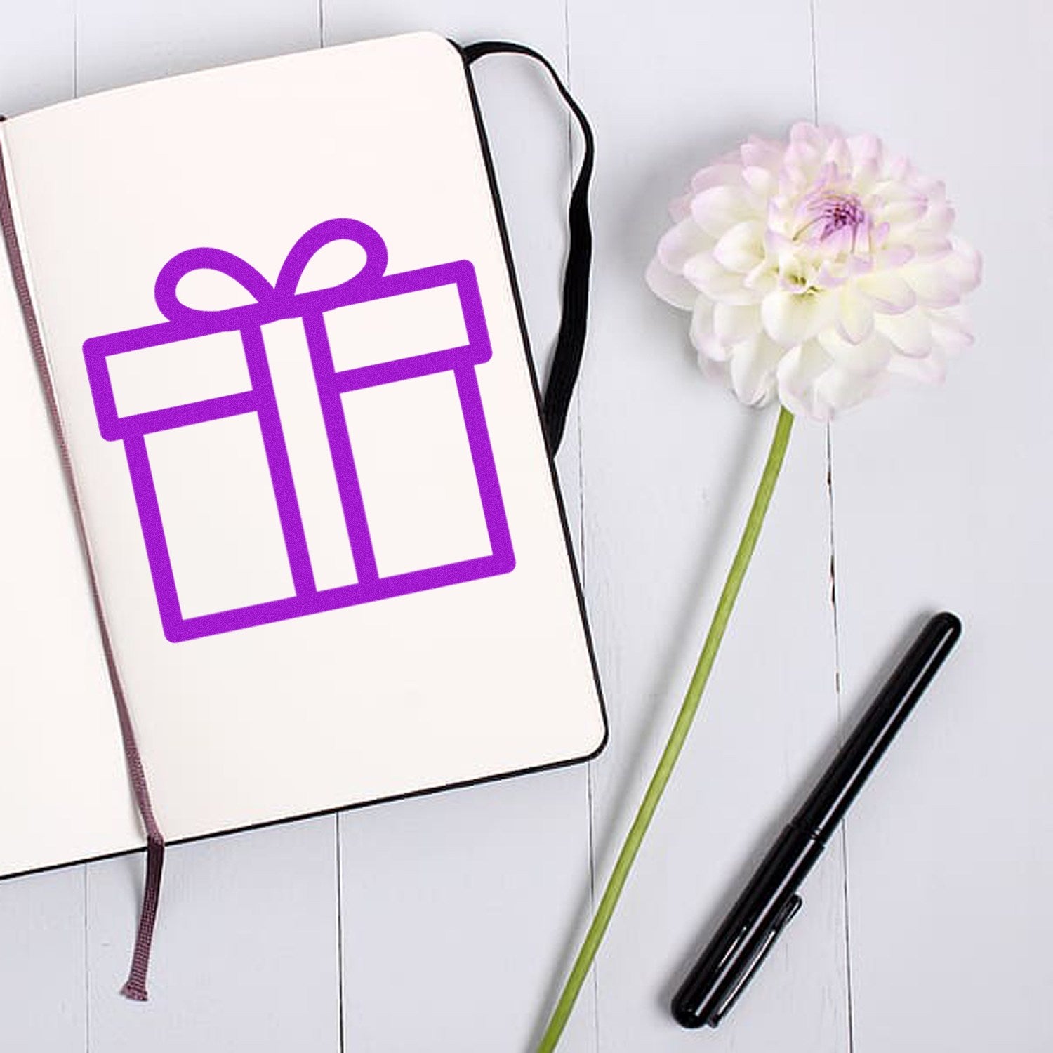 Wood Handle Gifts Galore Loyalty Points Rubber Stamp on a white notebook with a purple gift icon, next to a white flower and black pen on a light wooden surface.