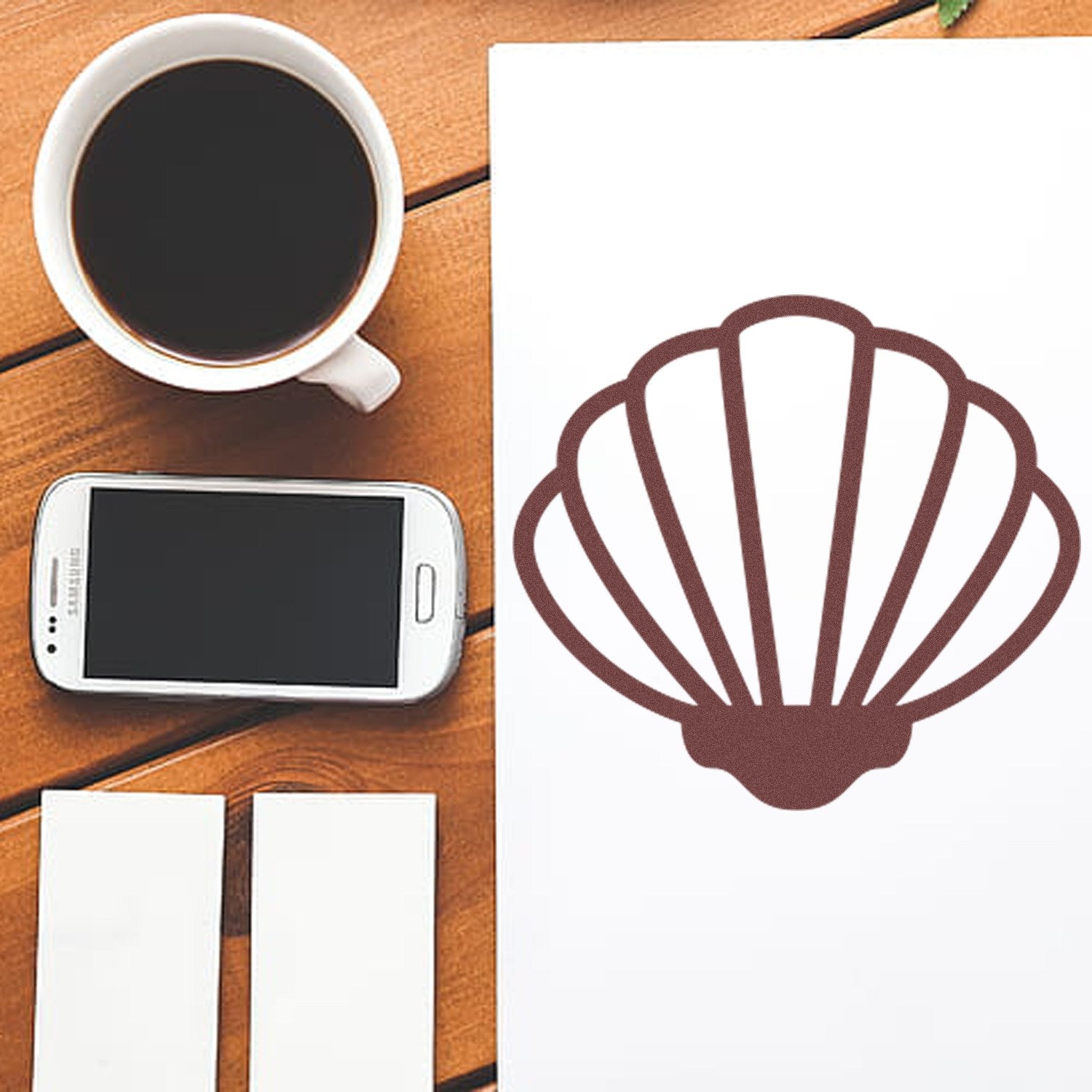 Self-Inking Beach Beauty Membership Stamp with a seashell design, shown on a wooden table beside a coffee cup, smartphone, and business cards.