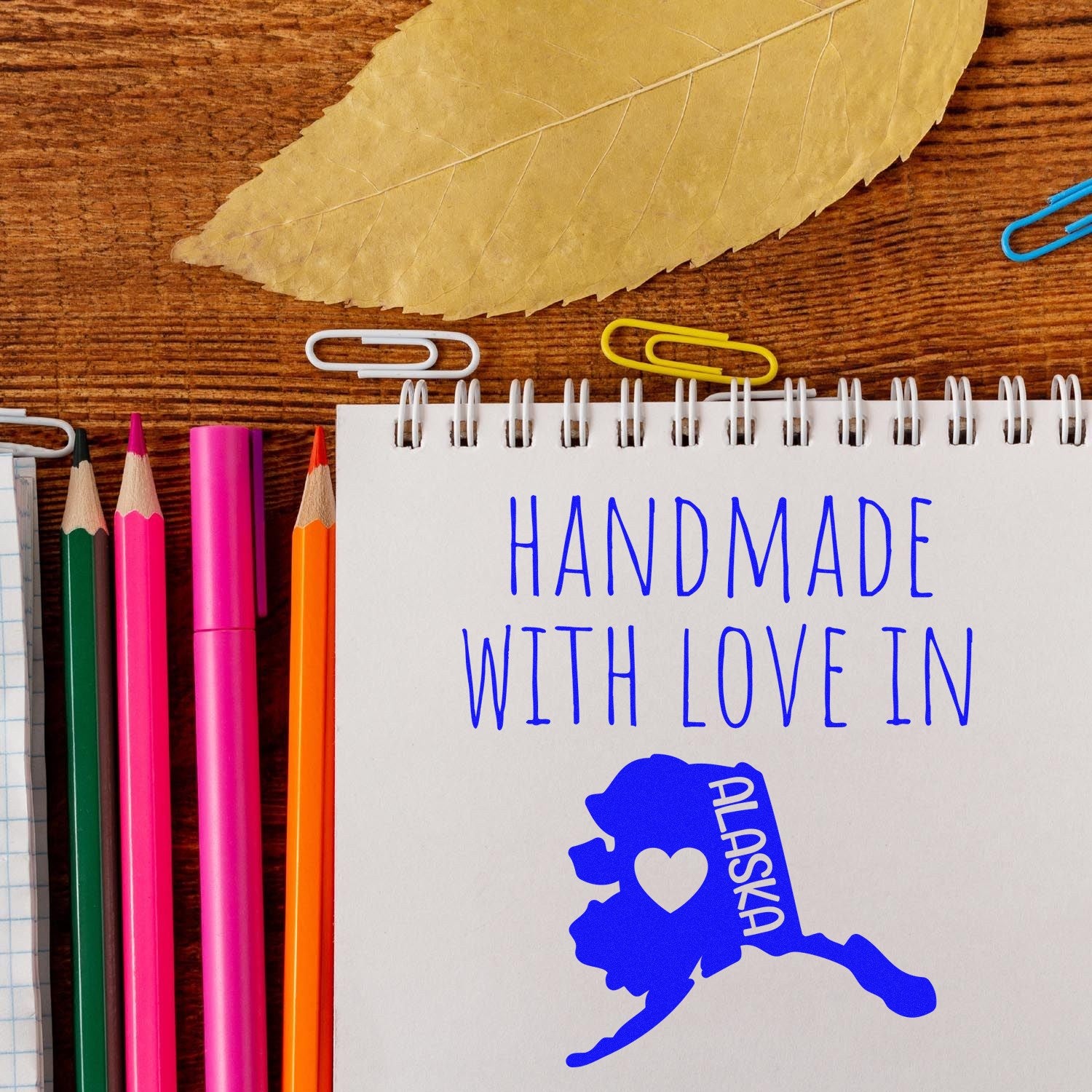 Wood Handle Alaska Handmade with Love Rubber Stamp on a wooden surface, surrounded by colorful pencils and paperclips, featuring a blue Alaska design with heart detail.