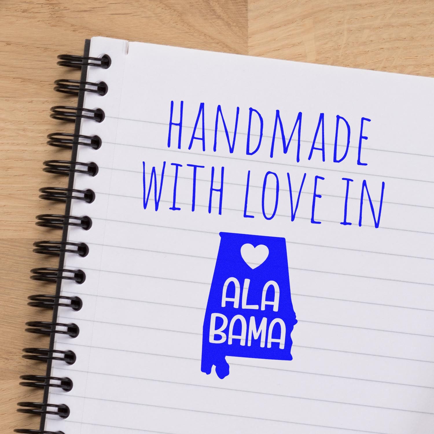 Handmade with Love in Alabama Slim Pre-Inked Stamp on a notebook, featuring blue text and a heart design within the Alabama state outline. Perfect for adding a personal touch to crafts.