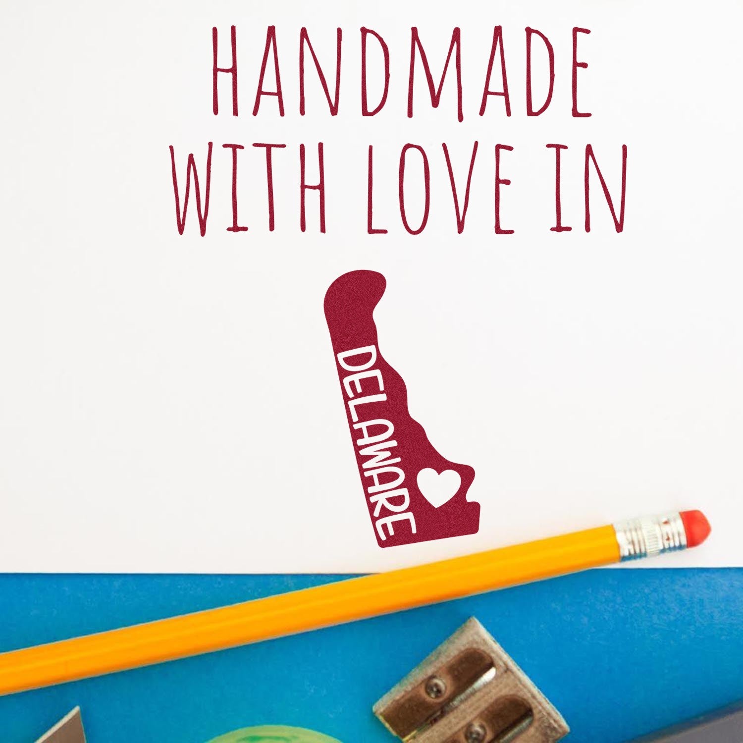 Wood Handle Delaware Handmade with Love Rubber Stamp featuring a Delaware state outline with 'Handmade with Love' text, shown with a pencil and sharpener on a blue surface.