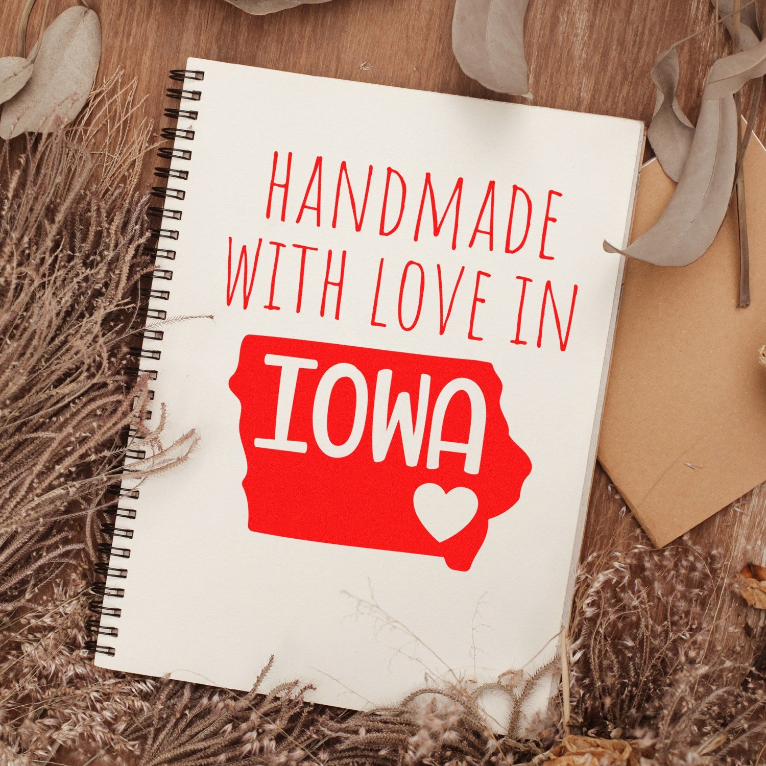 PSI Pre-Inked Handmade with Love in Iowa stamp on a spiral notebook, featuring red text and a heart on a map of Iowa, surrounded by dried leaves and a rustic background.