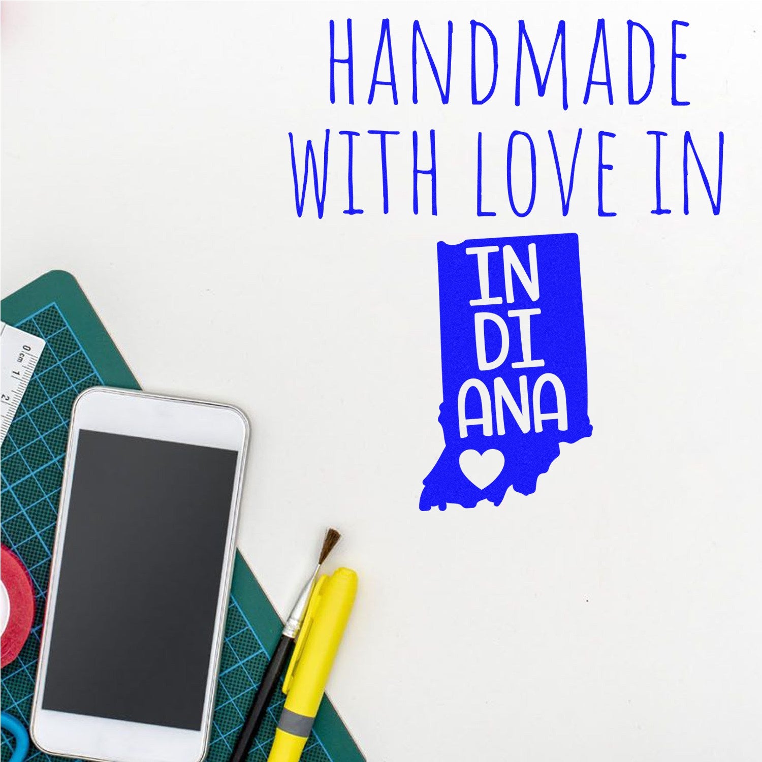 Wood Handle Indiana Handmade with Love Rubber Stamp on a crafting table with a smartphone, paintbrush, and yellow pen. Blue text and Indiana state outline with heart design.