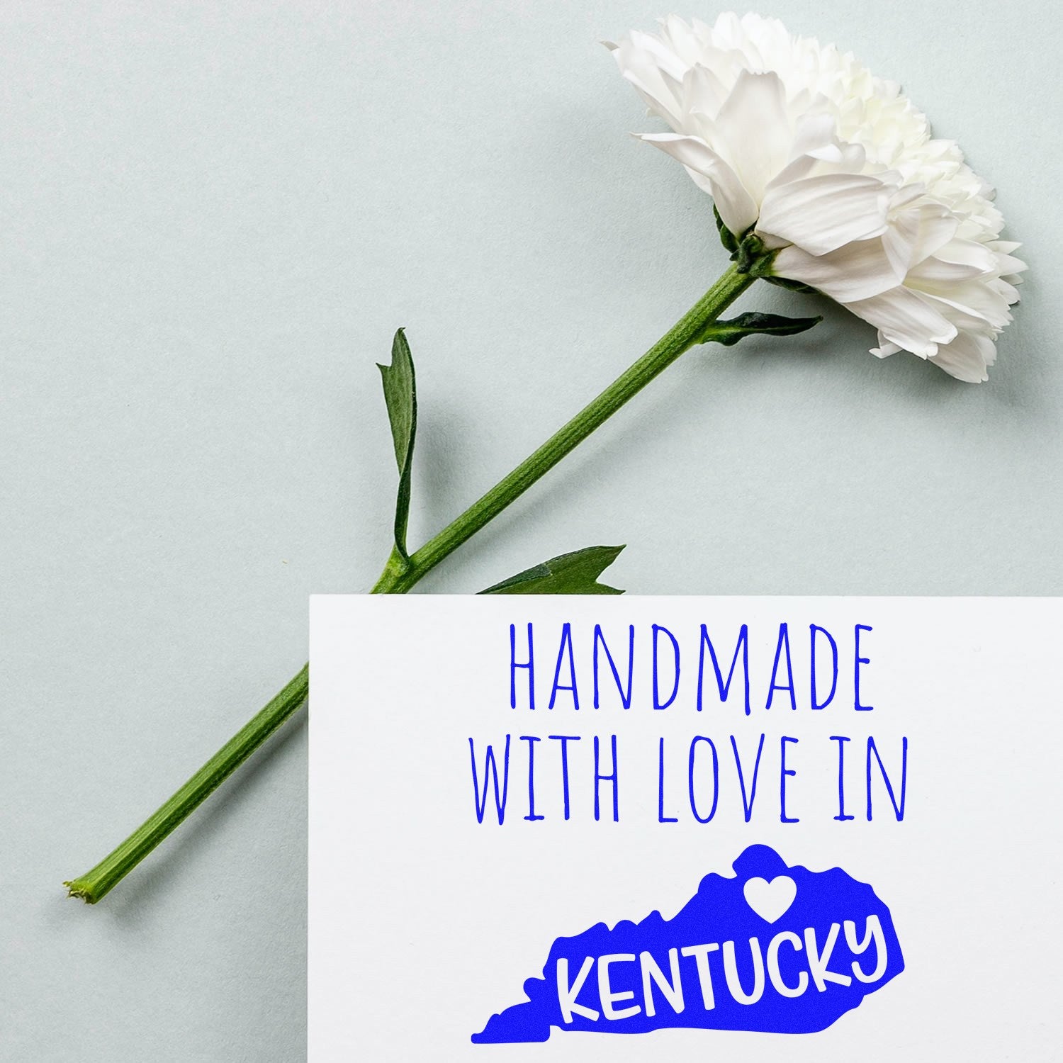 Self-Inking Kentucky Handmade with Love Stamp featuring a blue Kentucky state outline and heart, next to a white flower on a light background. Perfect for crafts and gifts.