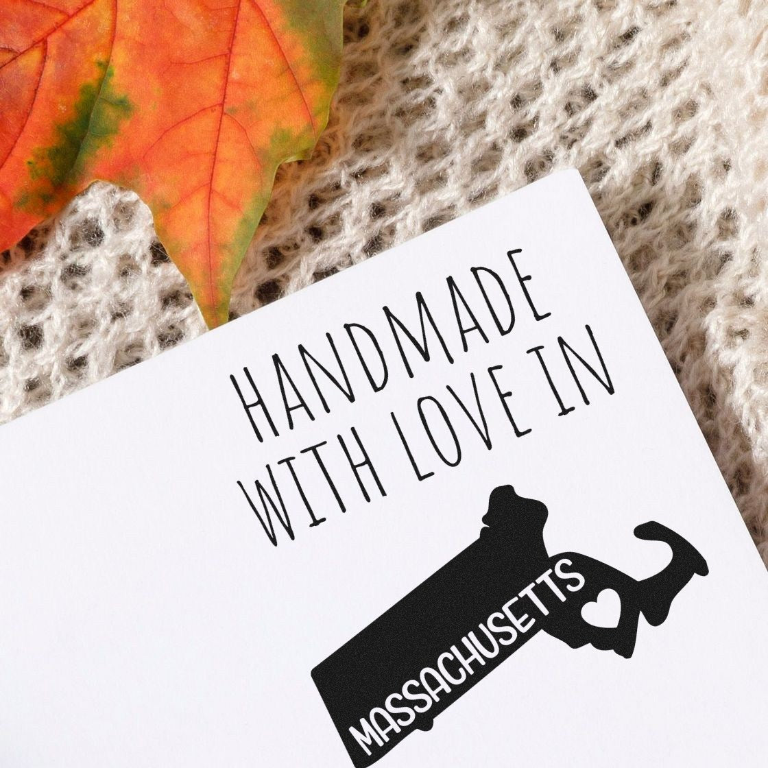 Handmade with Love in Massachusetts Slim Pre-Inked Stamp on a textured surface with a fall leaf, featuring a silhouette of Massachusetts with a heart.