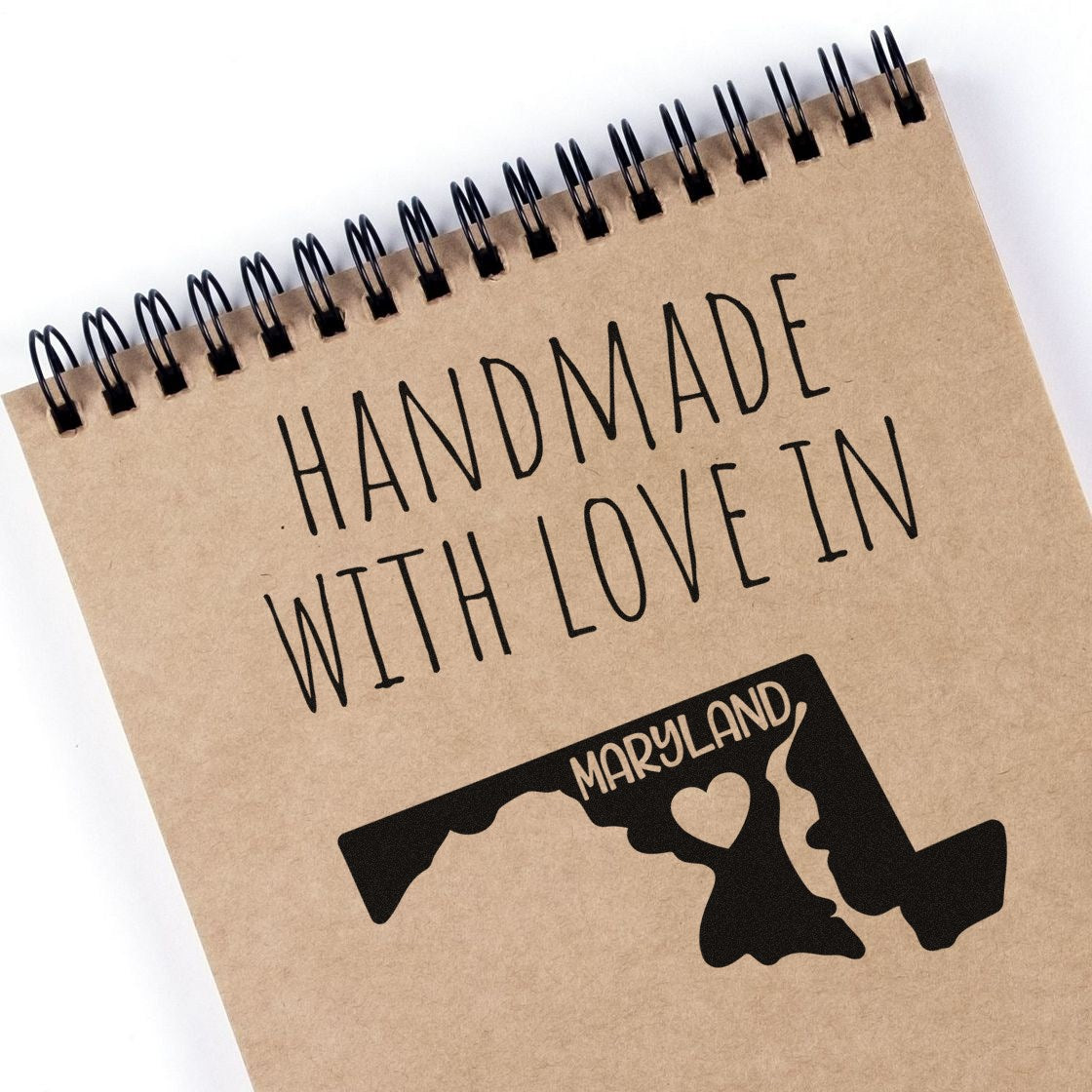 Self-Inking Maryland Handmade with Love Stamp on a notebook, featuring a black Maryland state outline with a heart. Perfect for crafts and gifts.