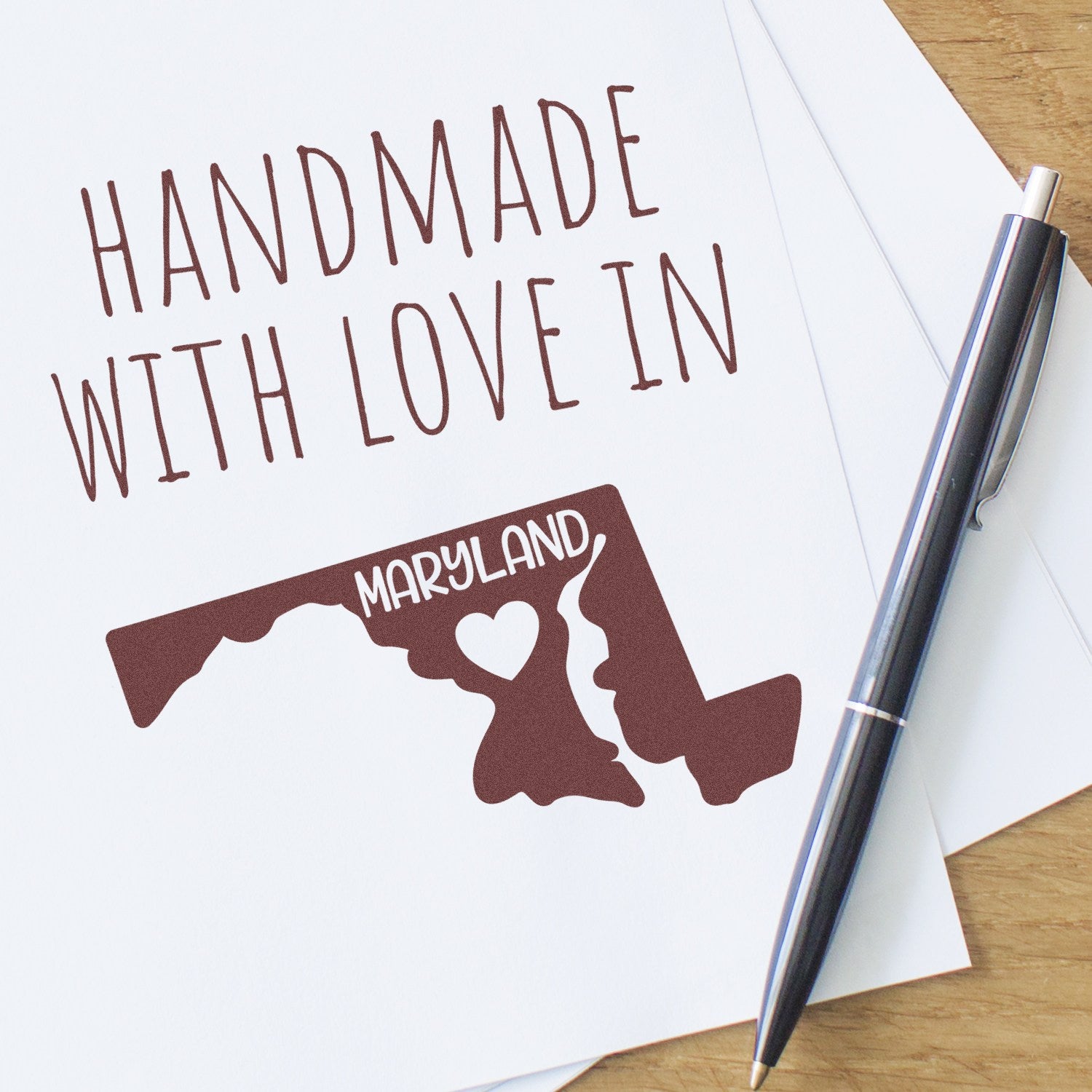 PSI Pre-Inked Handmade with Love in Maryland stamp on paper, featuring a Maryland map design with a heart, alongside a pen on a wooden surface.