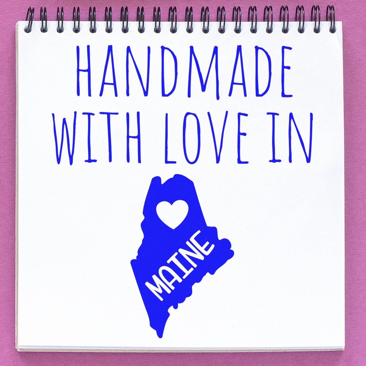 Handmade with Love in Maine Slim Pre-Inked Stamp on a notepad, featuring blue text and a heart-shaped Maine map design. Perfect for adding a personal touch to crafts and letters.