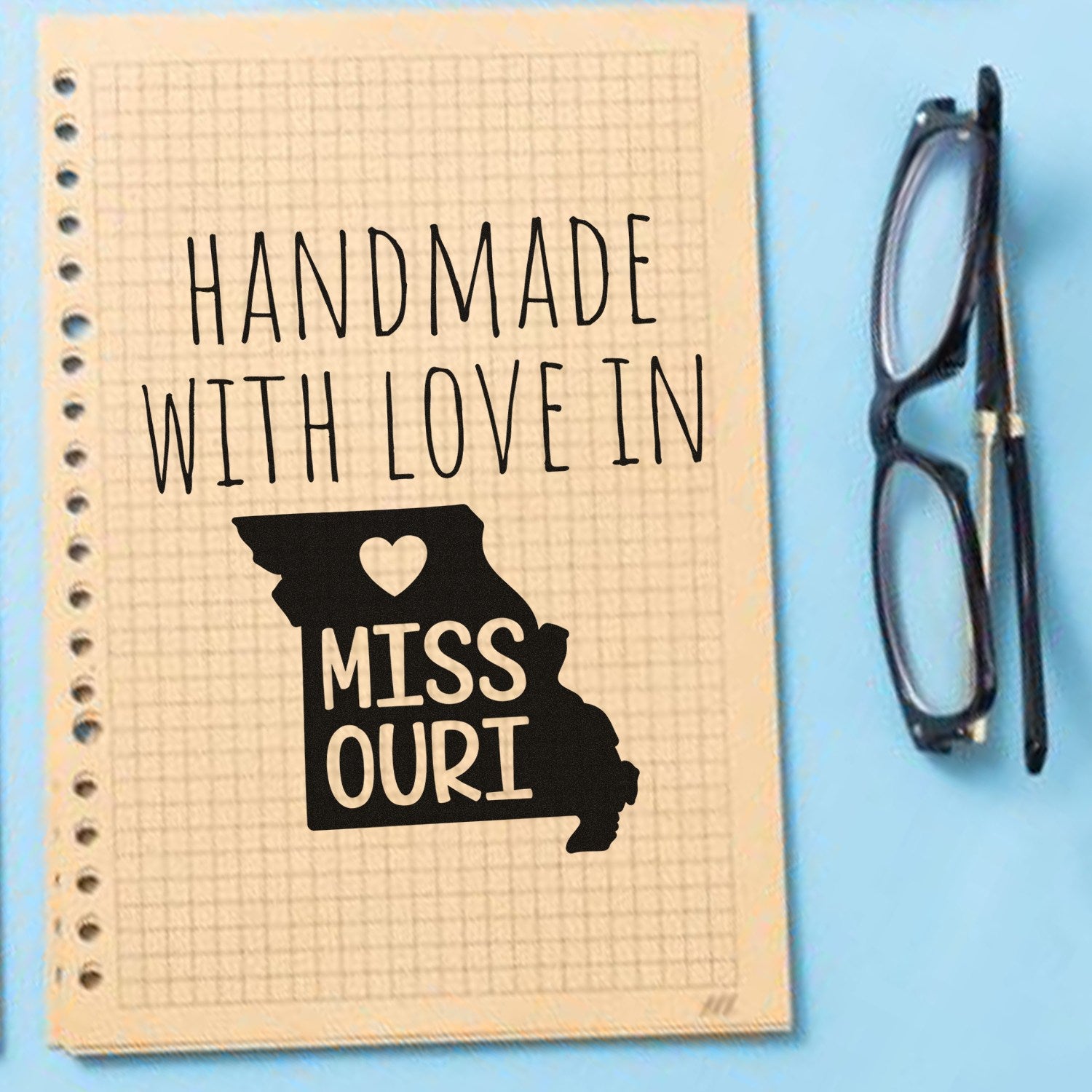 Handmade with Love in Missouri Slim Pre-Inked Stamp on grid paper, featuring a heart and Missouri state outline, next to black-rimmed glasses on a blue background.