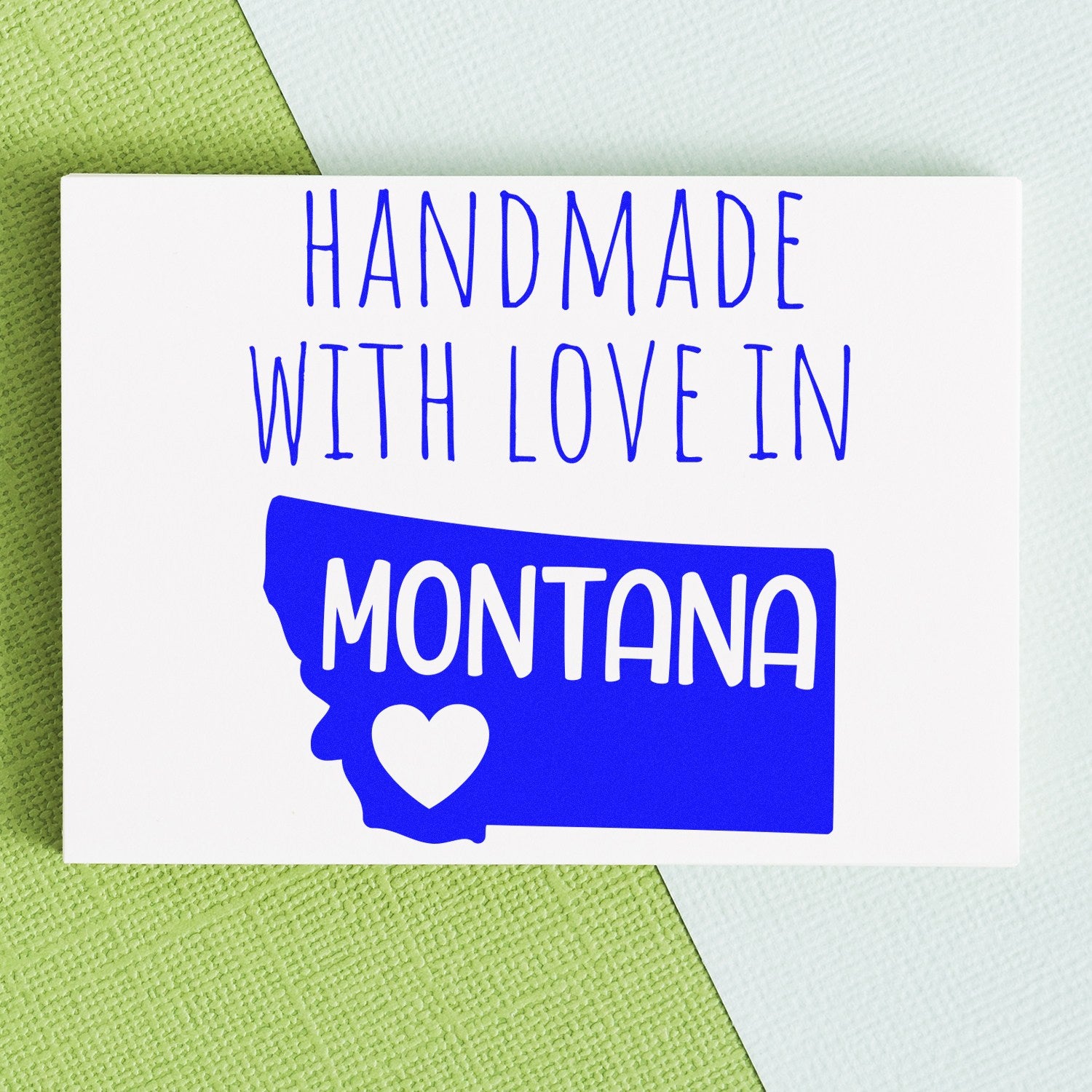 PSI Pre-Inked Handmade with Love in Montana stamp on a white card with blue text and a heart inside the Montana state outline, placed on a textured green and light blue background.