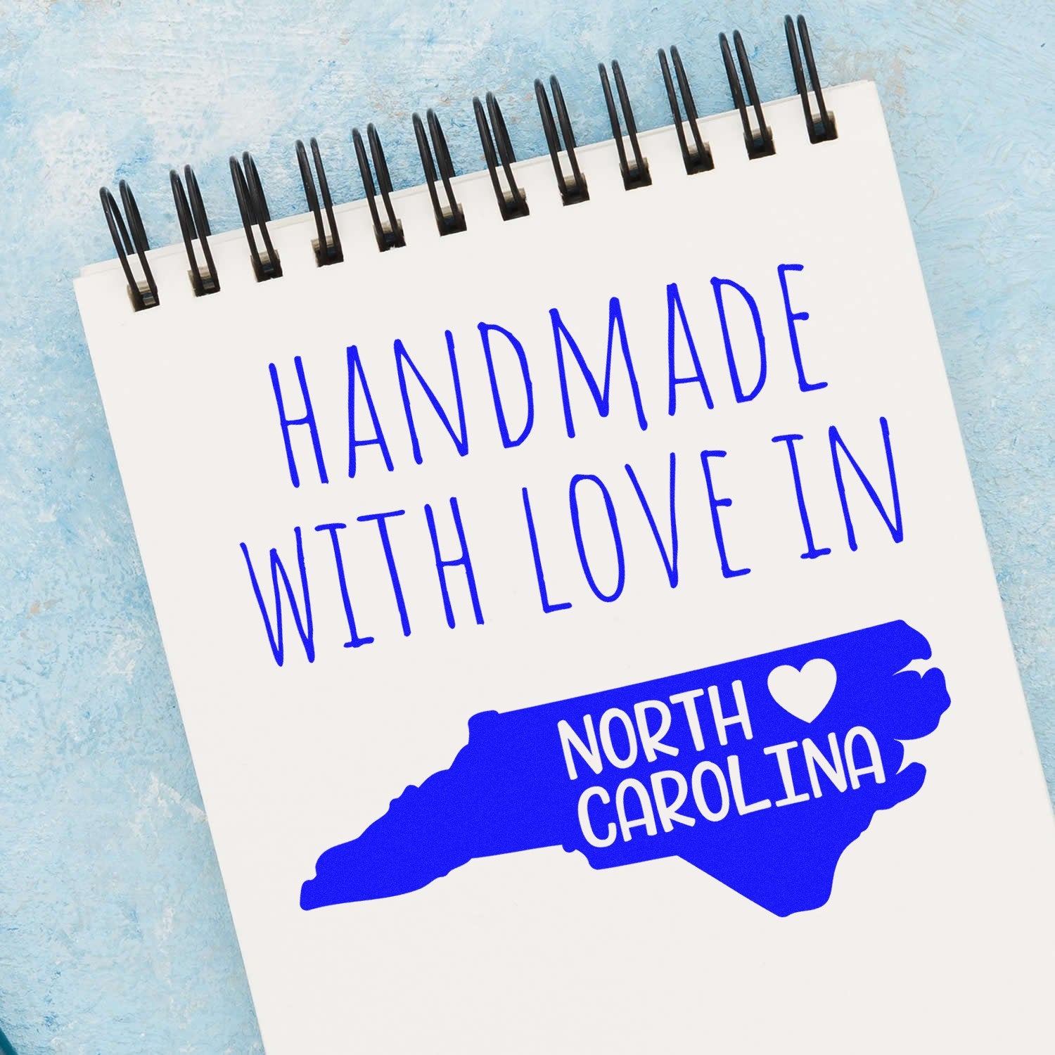 Handmade with Love in North Carolina Slim Pre-Inked Stamp on a notepad, featuring blue text and a heart within the state outline, showcasing its artisanal design.