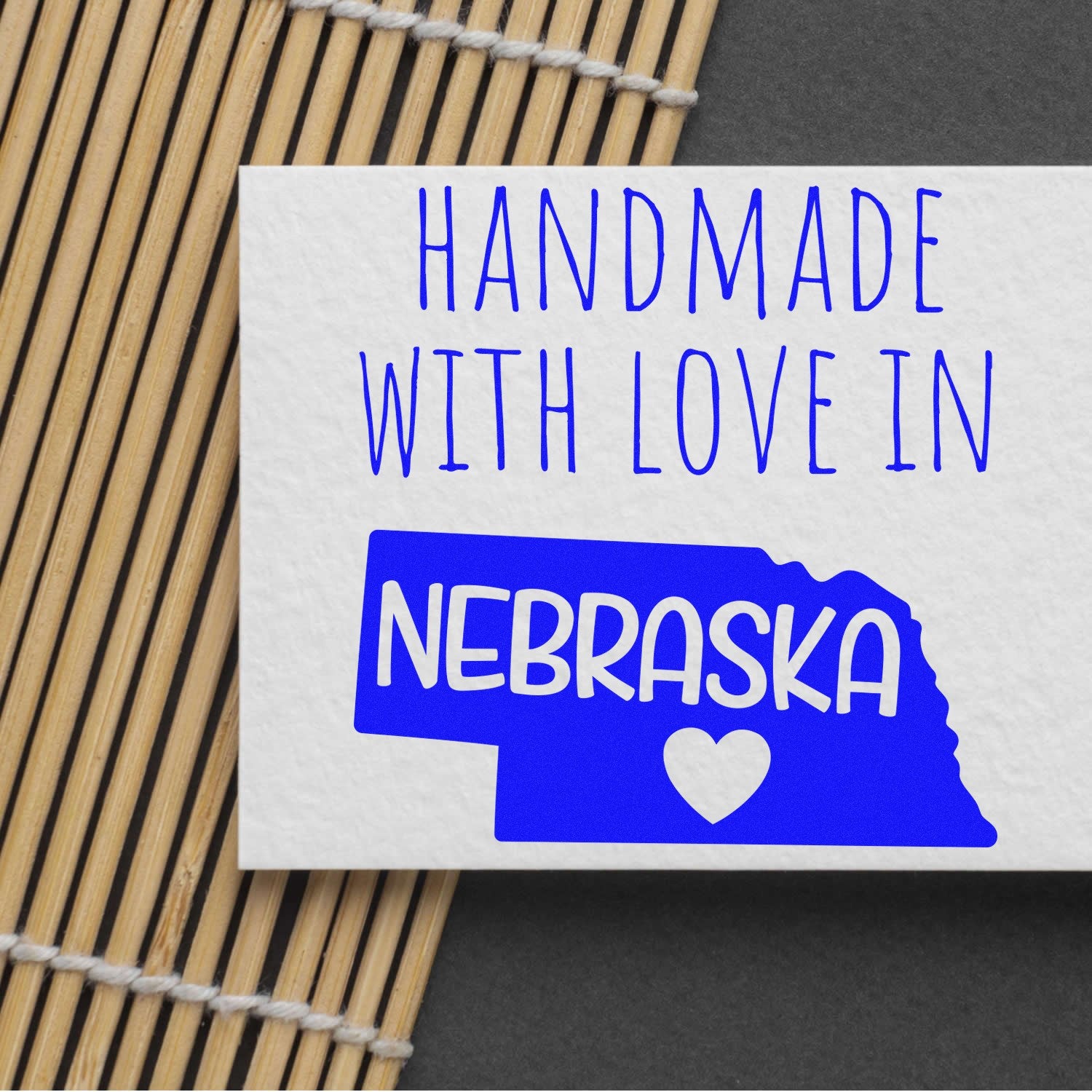 Wood Handle Nebraska Handmade with Love Rubber Stamp on a card, featuring a blue Nebraska state outline with a heart, placed on a bamboo mat background.