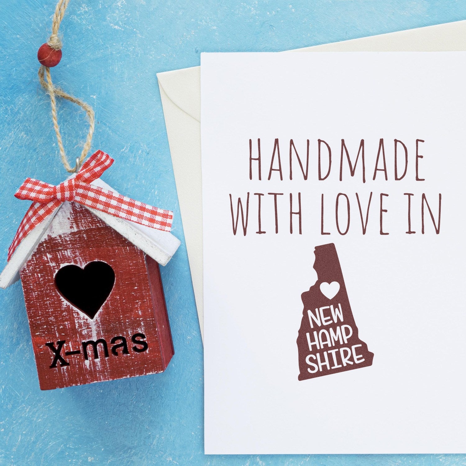 Wood Handle New Hampshire Handmade with Love Rubber Stamp on a blue background, next to a card and a small decorative house with a heart and X-mas text.