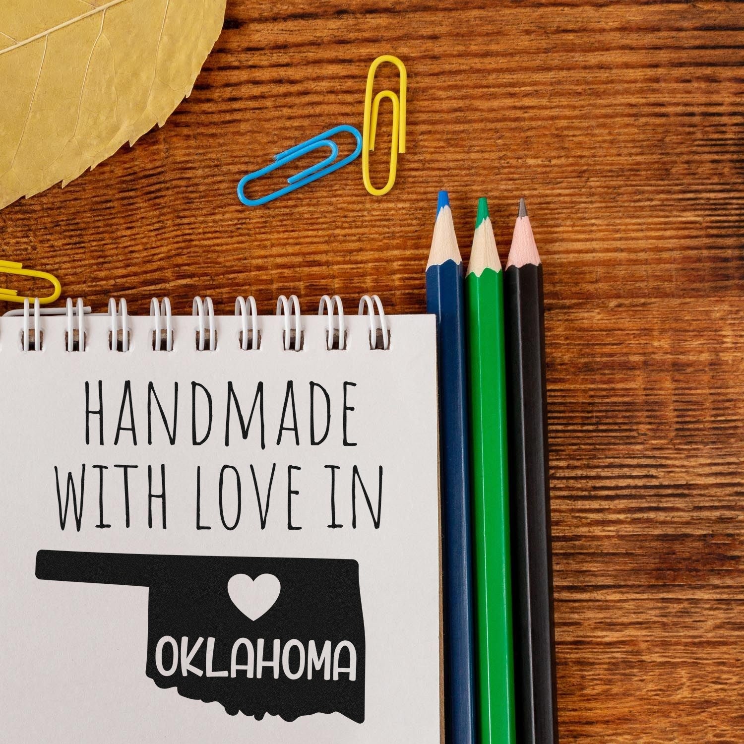 Self-Inking Oklahoma Handmade with Love Stamp on notepad, featuring a heart and state outline, with colorful pencils and paperclips on a wooden surface.