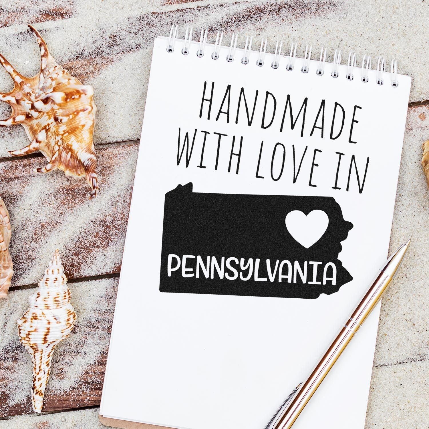 Wood Handle Pennsylvania Handmade with Love Rubber Stamp on a notebook, featuring a heart and state outline, surrounded by seashells on a sandy surface.