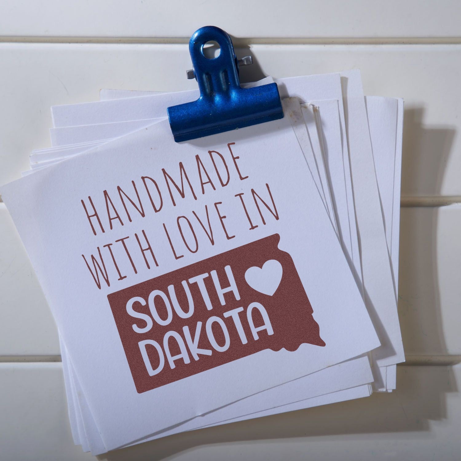 Handmade with Love in South Dakota Slim Pre-Inked Stamp on white paper, featuring a heart and state outline, clipped with a blue binder on a white background.