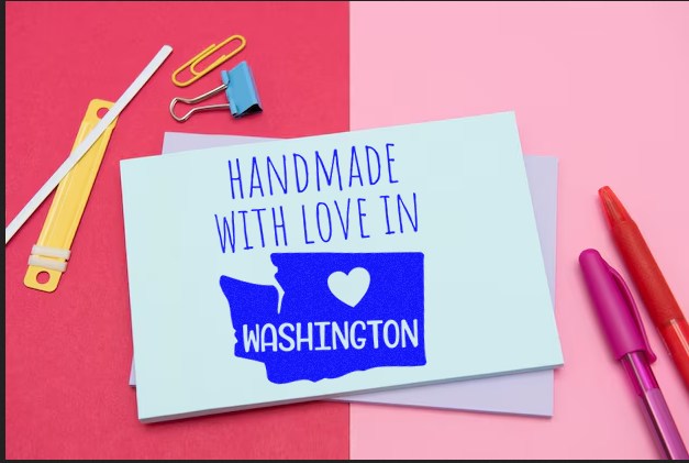 Self-Inking Washington Handmade with Love Stamp on a card, featuring a blue map of Washington with a heart, surrounded by stationery on a pink and red background.