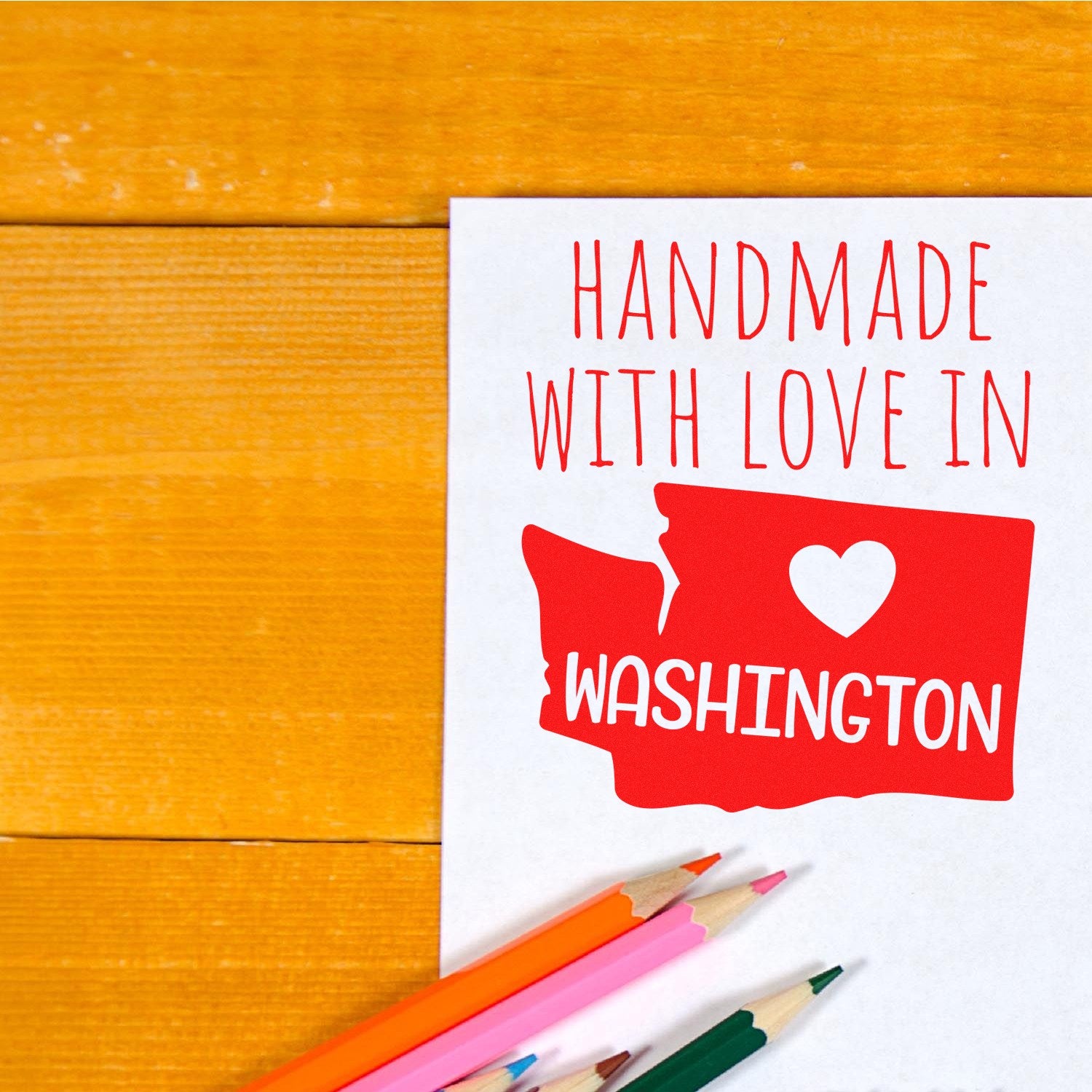 Handmade with Love in Washington Slim Pre-Inked Stamp on paper, featuring red text and a heart over a Washington state outline, with colored pencils nearby on a wooden surface.