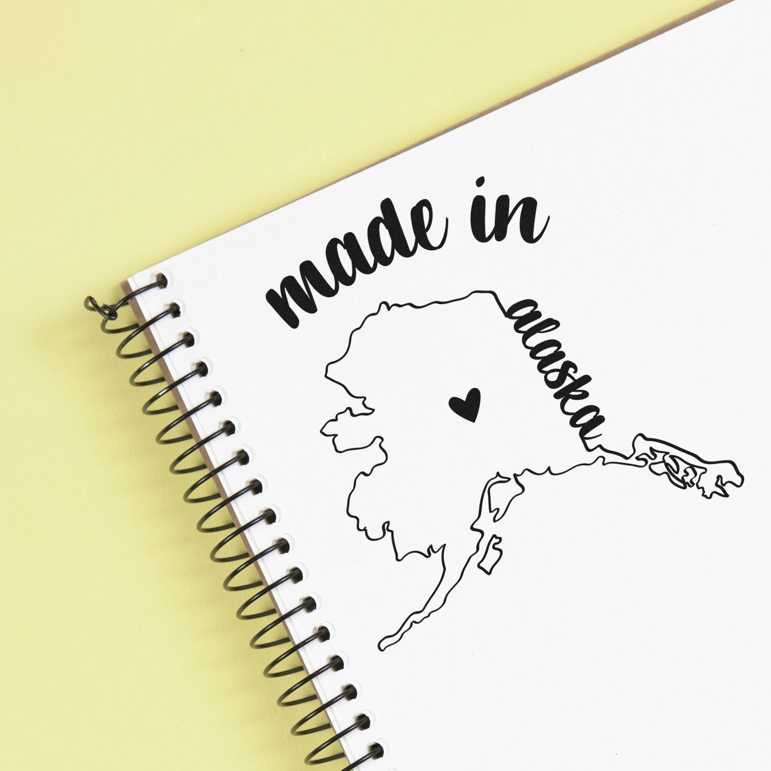 Made with Love in Alaska Rubber Stamp on a spiral notebook, featuring a heart and Alaska map design, perfect for adding a personal touch to crafts and stationery.