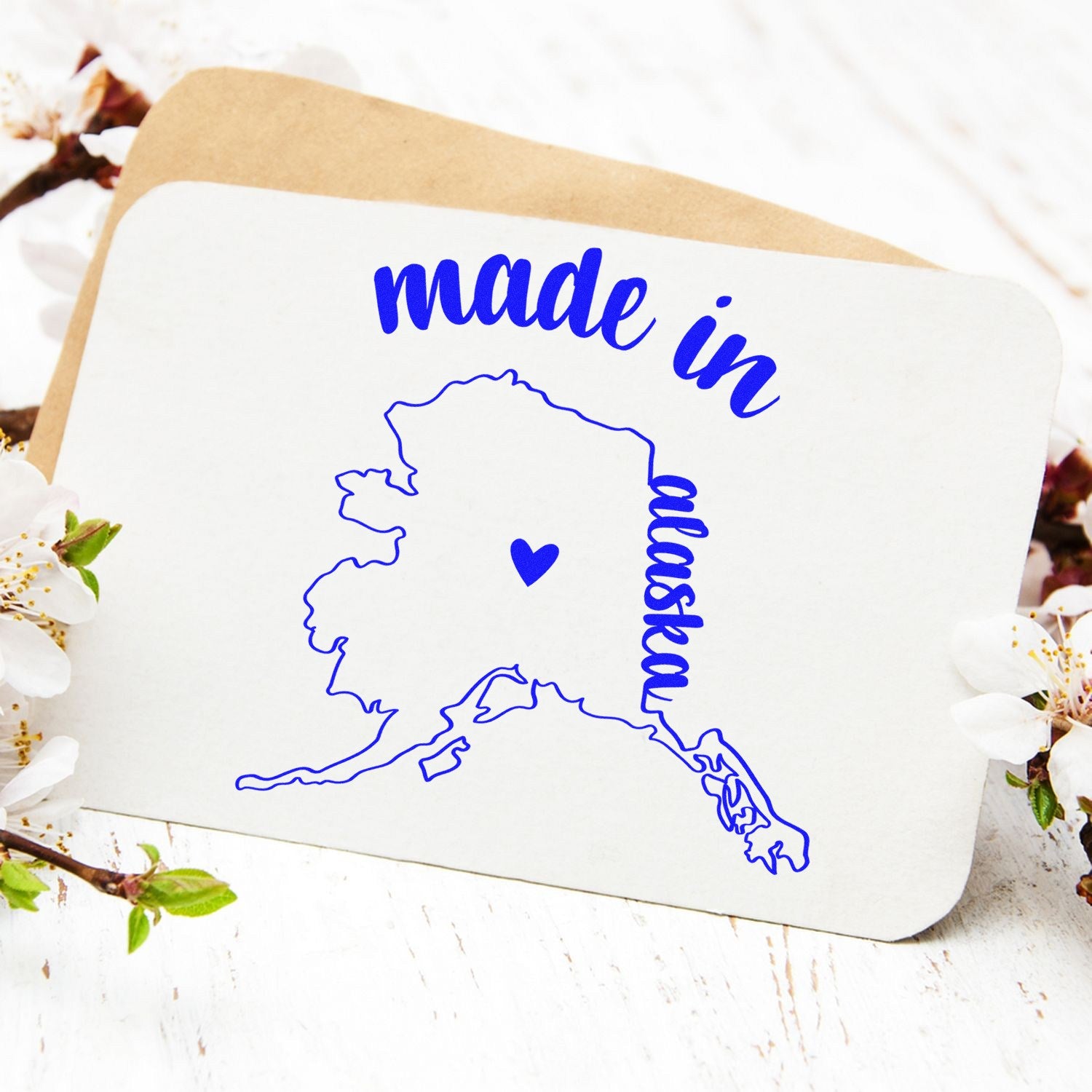 Made with Love in Alaska Rubber Stamp featuring a blue outline of Alaska with a heart, surrounded by flowers on a white background.