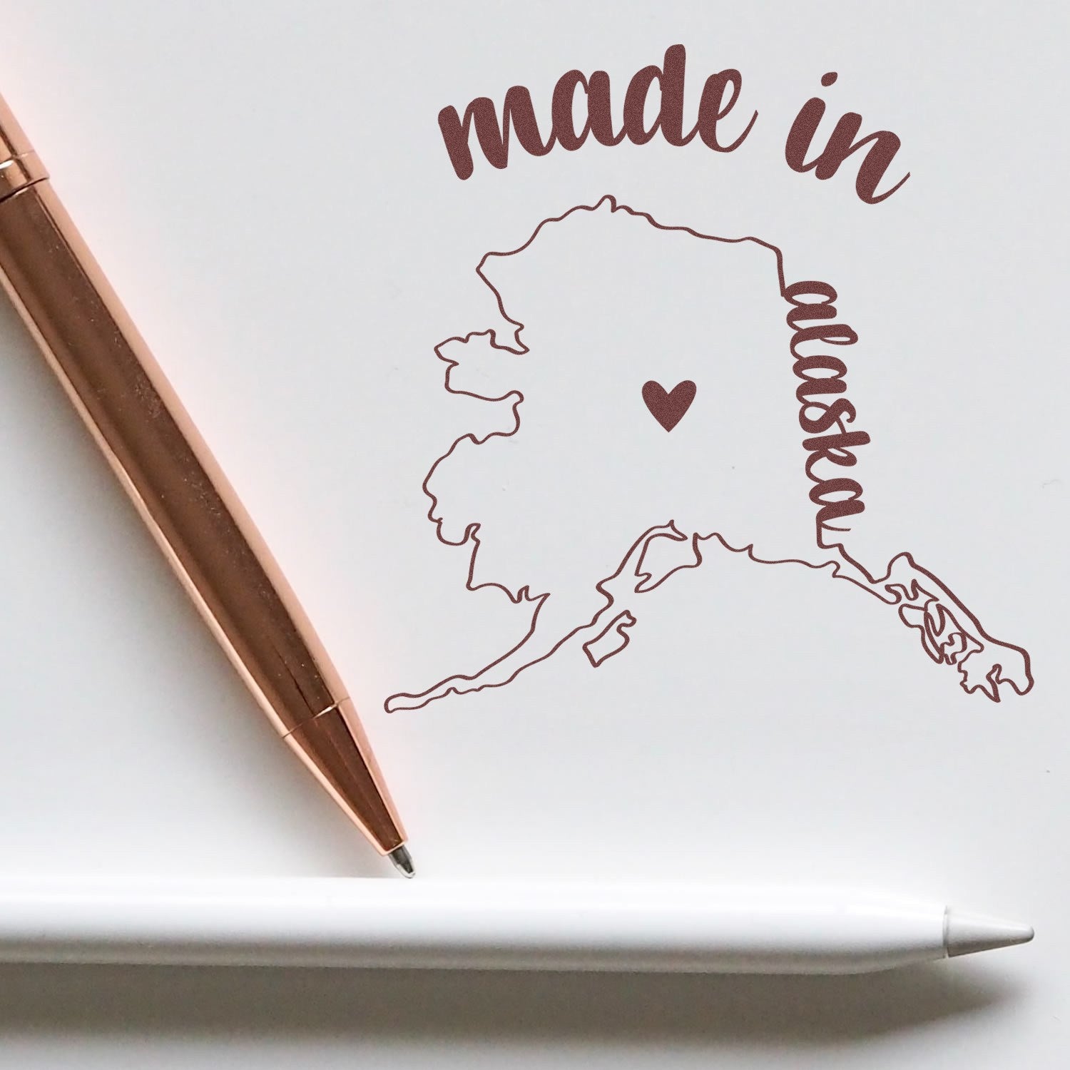 Self-Inking Handmade with Love in Alaska Stamp featuring a map of Alaska with a heart, alongside a rose gold pen on white paper.