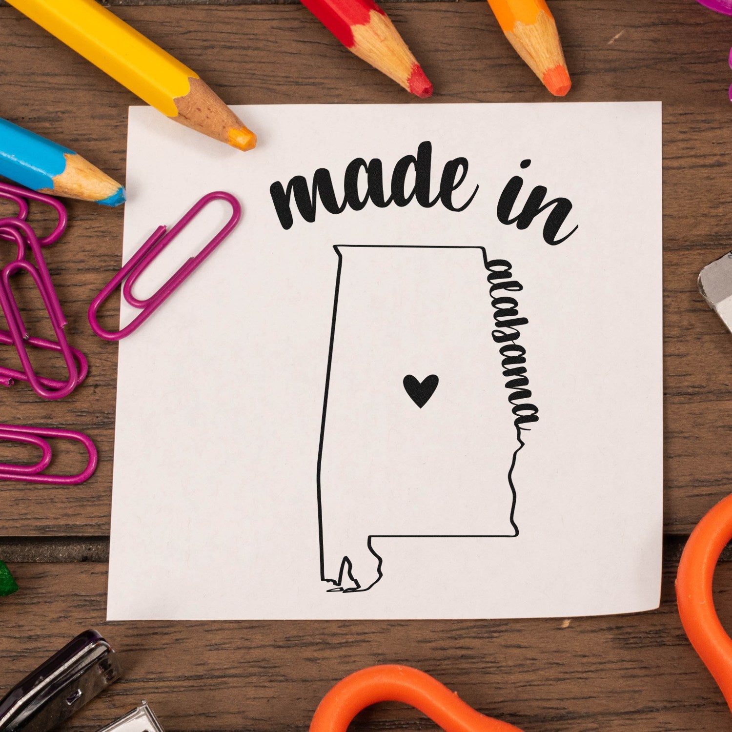 Made in Alabama Stamp Pre-Inked featuring a heart inside the state outline, surrounded by colorful pencils and paperclips on a wooden surface.