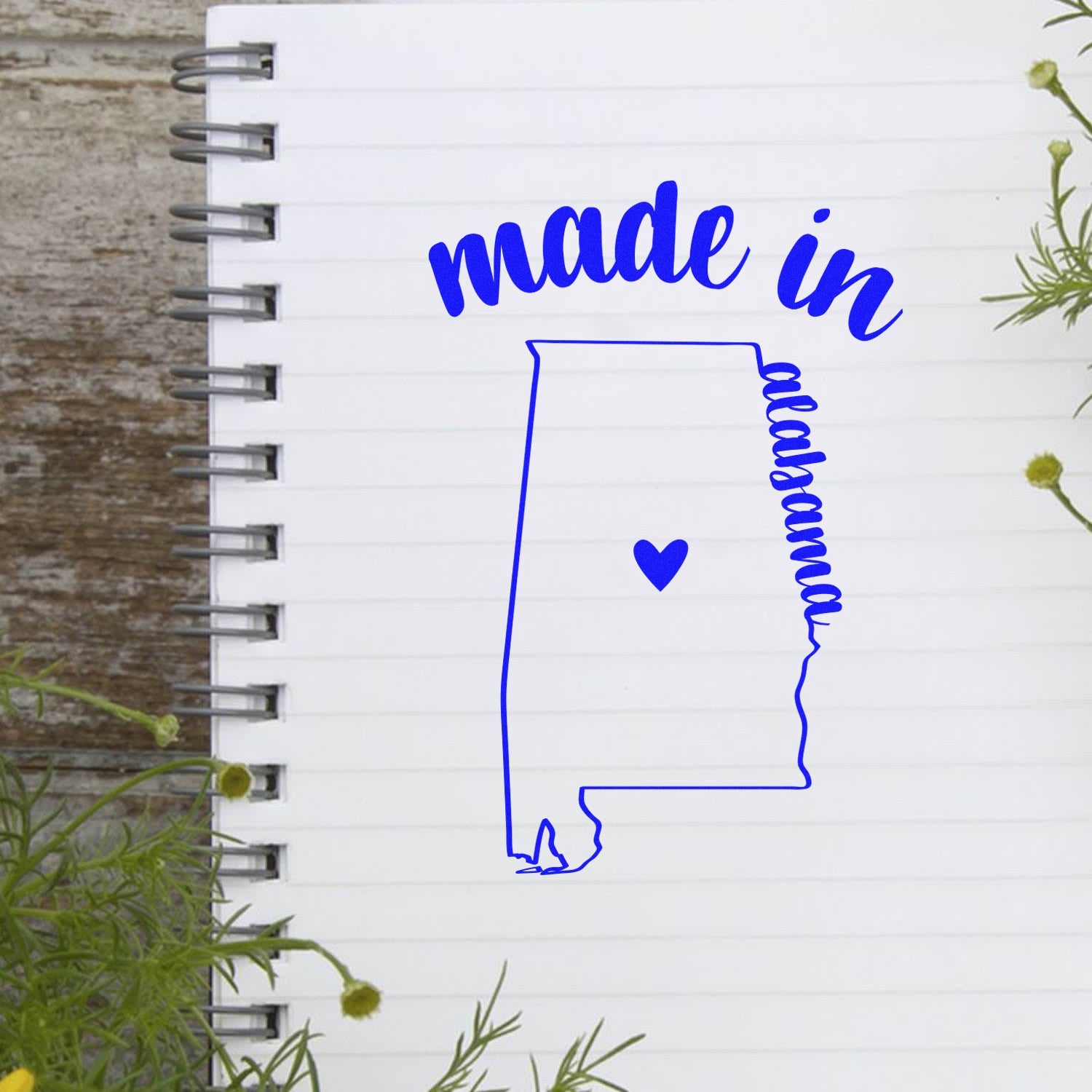 Made with Love in Alabama Rubber Stamp featuring a blue outline of Alabama with a heart, placed on a notebook. Perfect for crafts and personalized projects.