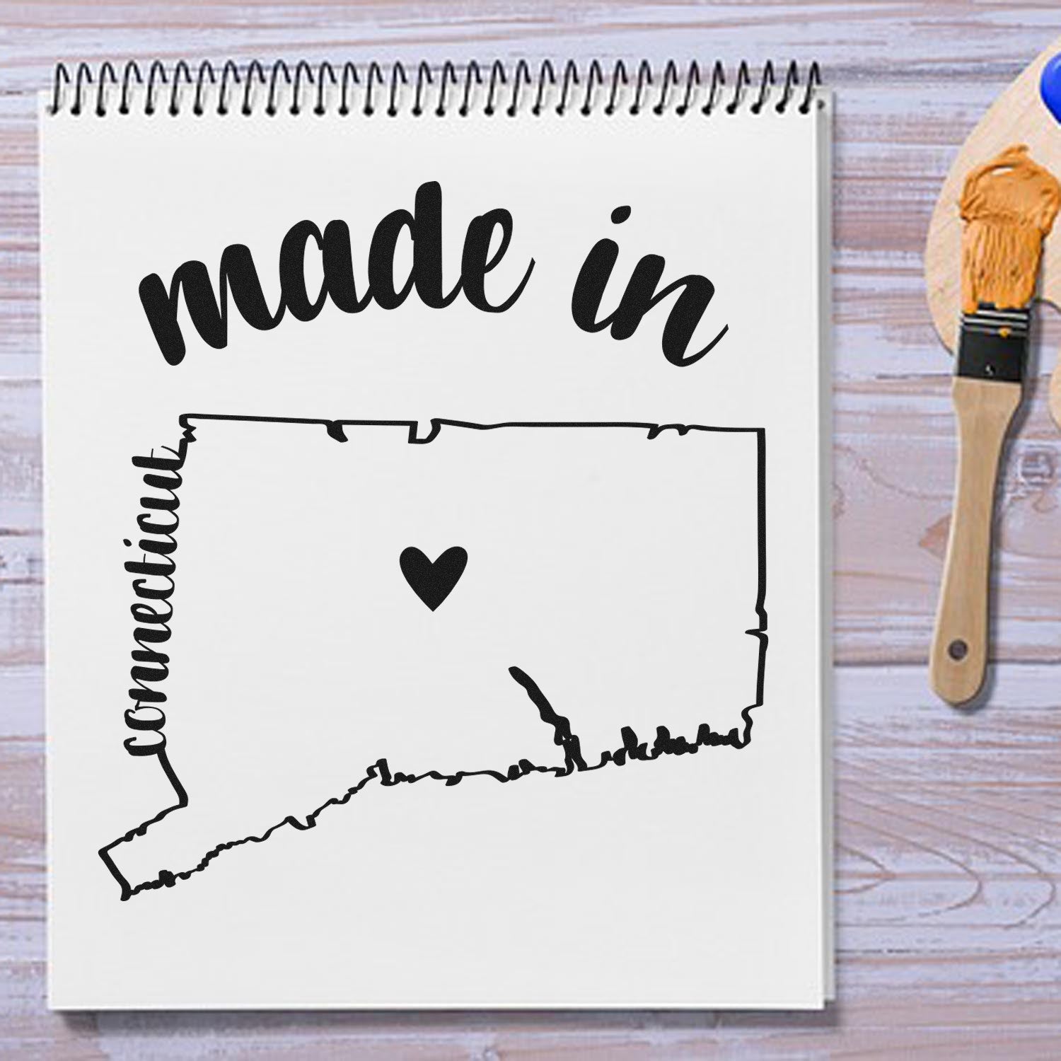 Made with Love in Connecticut Rubber Stamp featuring a heart inside a Connecticut outline, displayed on a notepad with a paintbrush on a wooden surface.