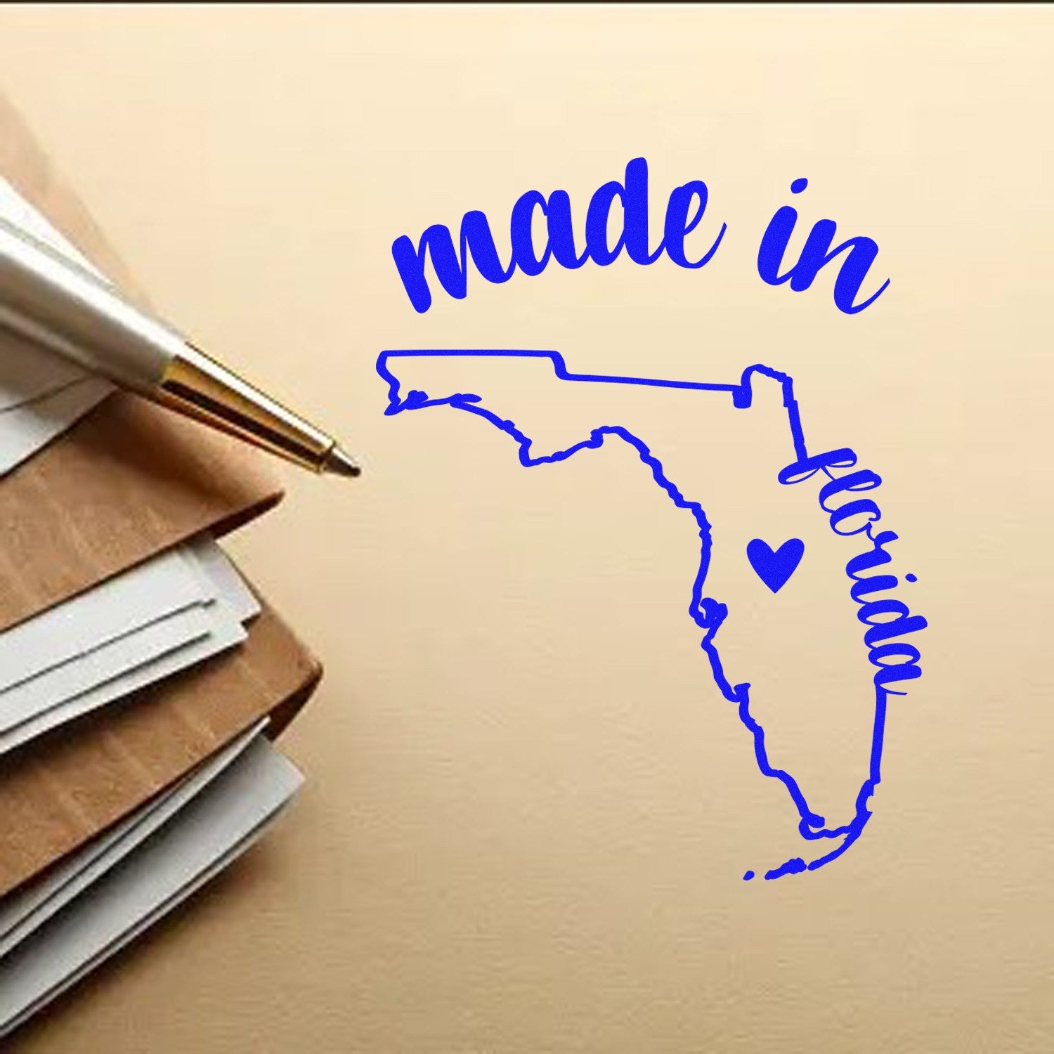 Made in Florida Stamp Pre-Inked featuring a blue outline of Florida with 'made in' and 'Florida' text, alongside a heart symbol, placed on a stack of papers with a pen.