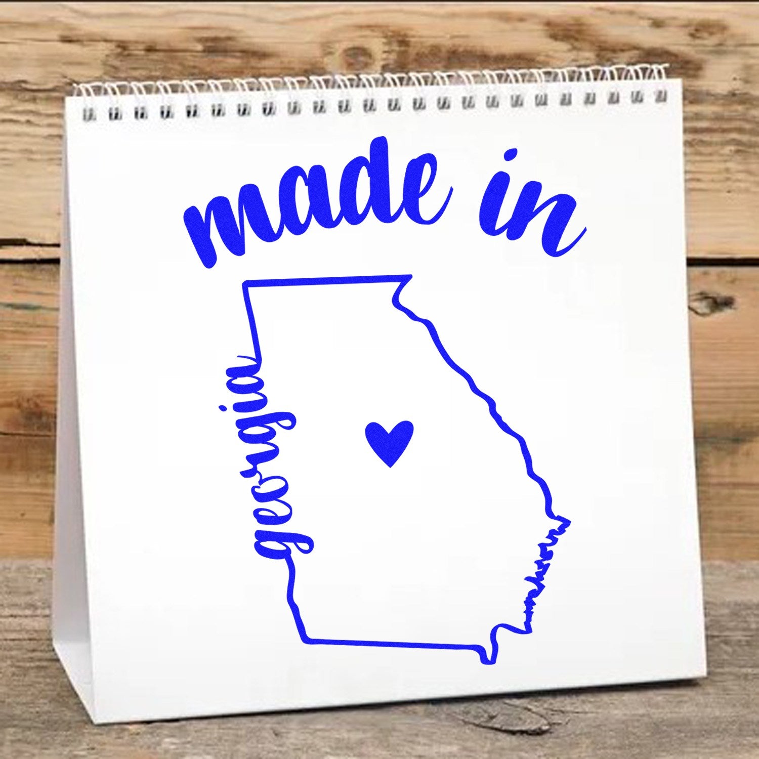 Image of Slim Pre-Inked Stamp Georgia Made in Stamp, featuring a blue outline of Georgia with made in text and a heart, on a spiral-bound notepad against a wooden background.