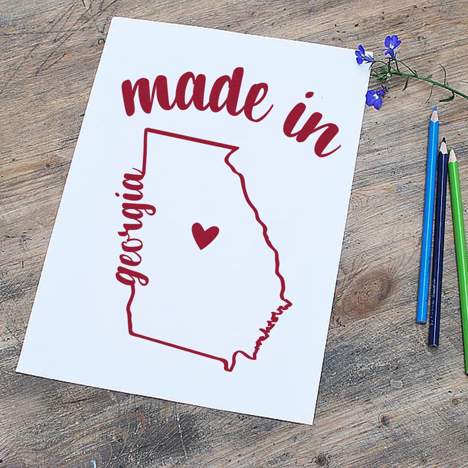 Self-Inking Handmade with Love in Georgia Stamp on paper, featuring a red outline of Georgia with a heart, surrounded by colored pencils on a wooden surface.