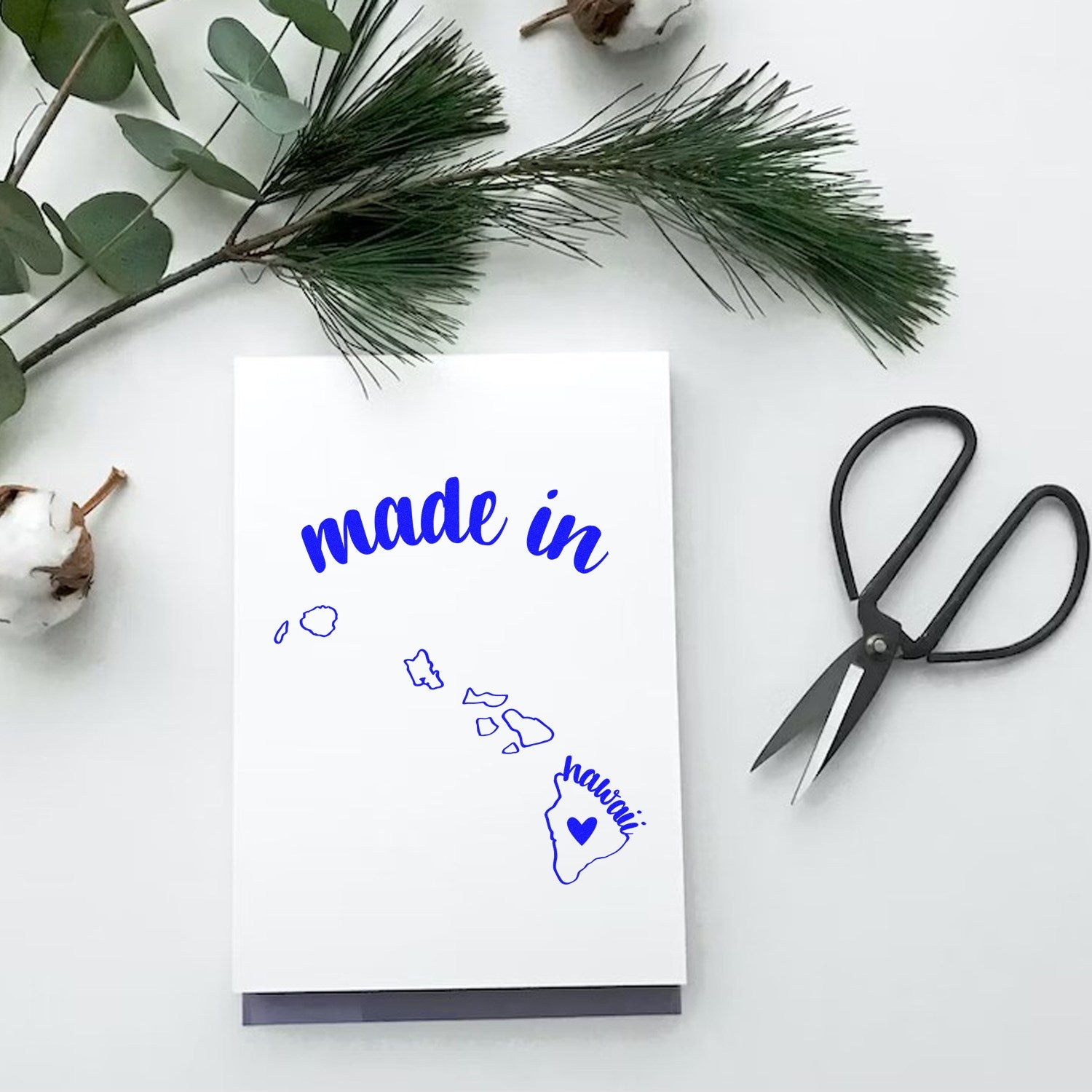 Made with Love in Hawaii Rubber Stamp on white paper, featuring blue text and a heart over Hawaii, surrounded by greenery and scissors.