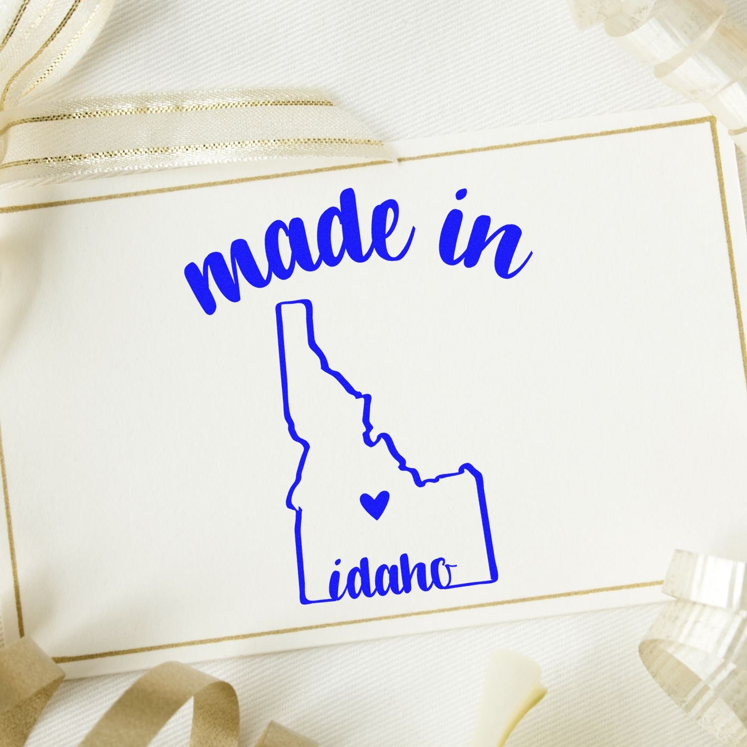 Made with Love in Idaho Rubber Stamp on a card with blue text and Idaho state outline, surrounded by ribbon. Perfect for crafts and gifts.