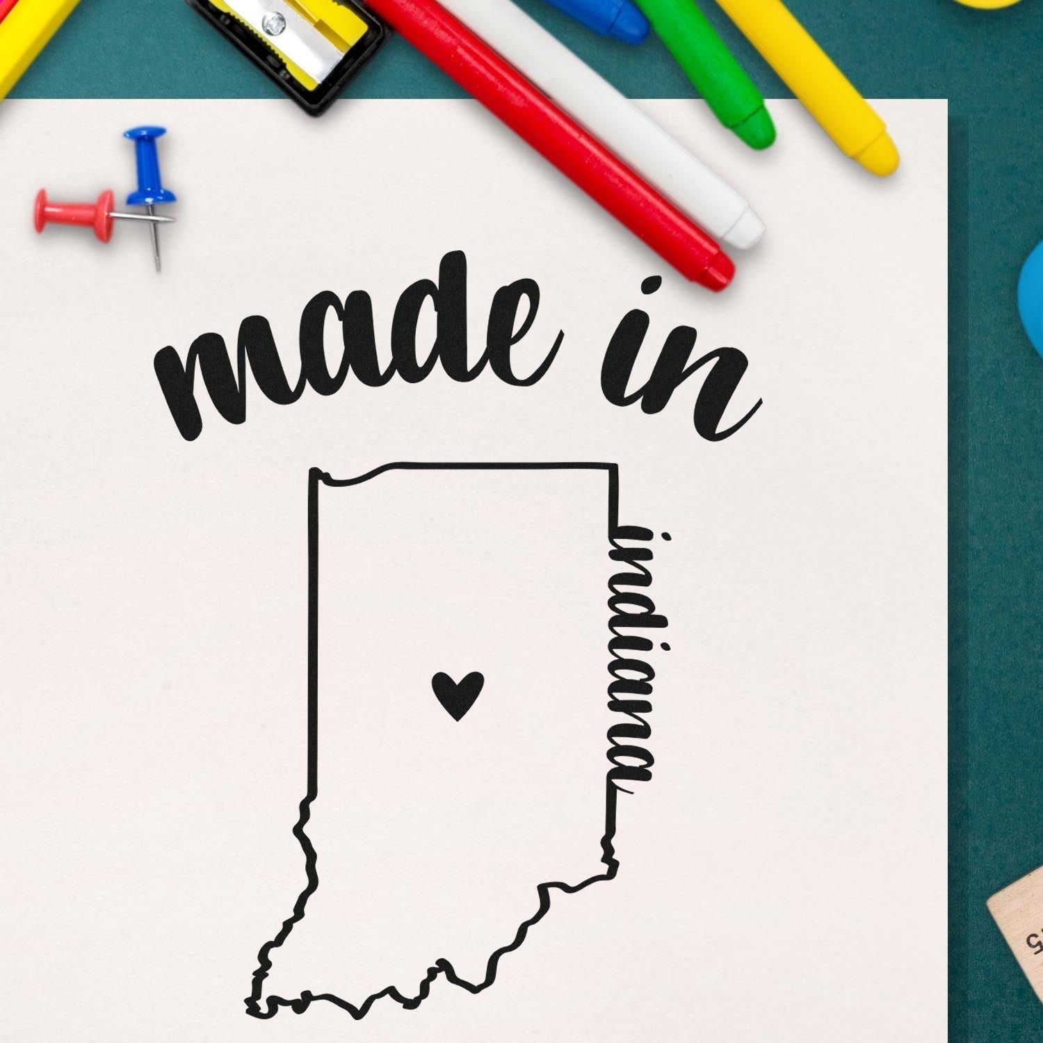 Made in Indiana Stamp Pre-Inked featuring a heart inside the Indiana state outline, surrounded by colorful office supplies on a green background.