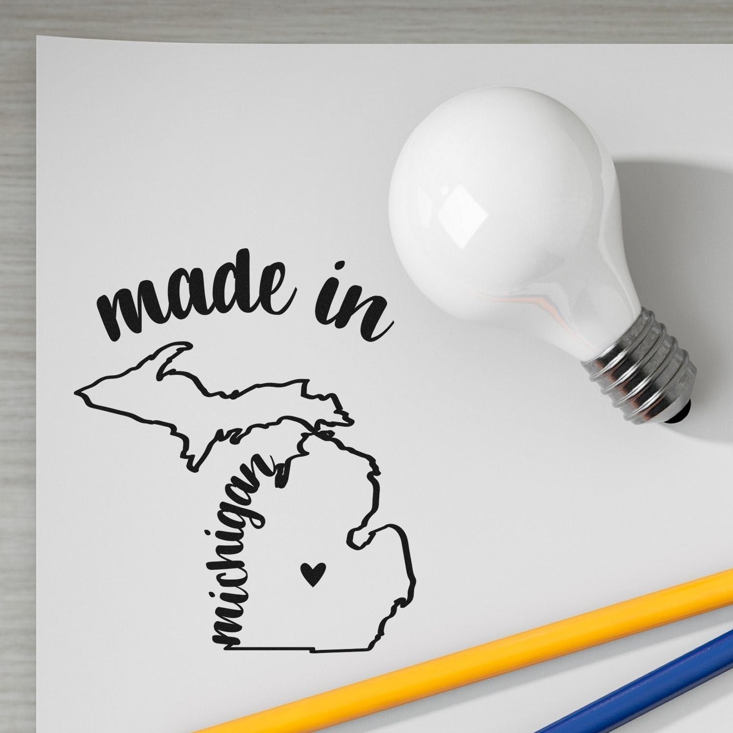 Image of a Slim Pre-Inked Stamp Michigan Made in Stamp, featuring a map of Michigan with made in text, next to a light bulb and pencils on a white surface. Perfect for crafts and branding.