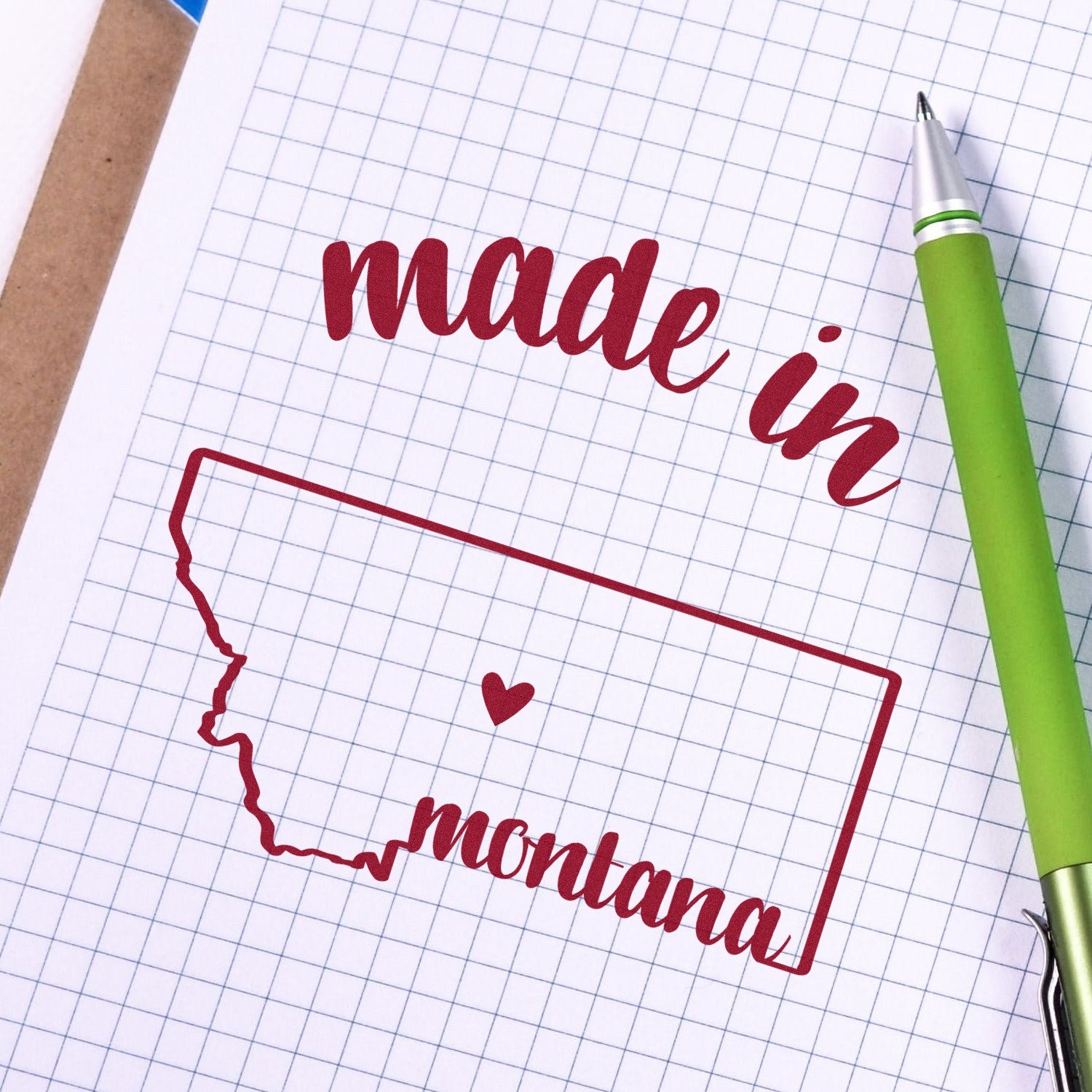 Self-Inking Handmade with Love in Montana Stamp featuring a red outline of Montana with 'made in montana' text and heart, shown on graph paper with a green pen.