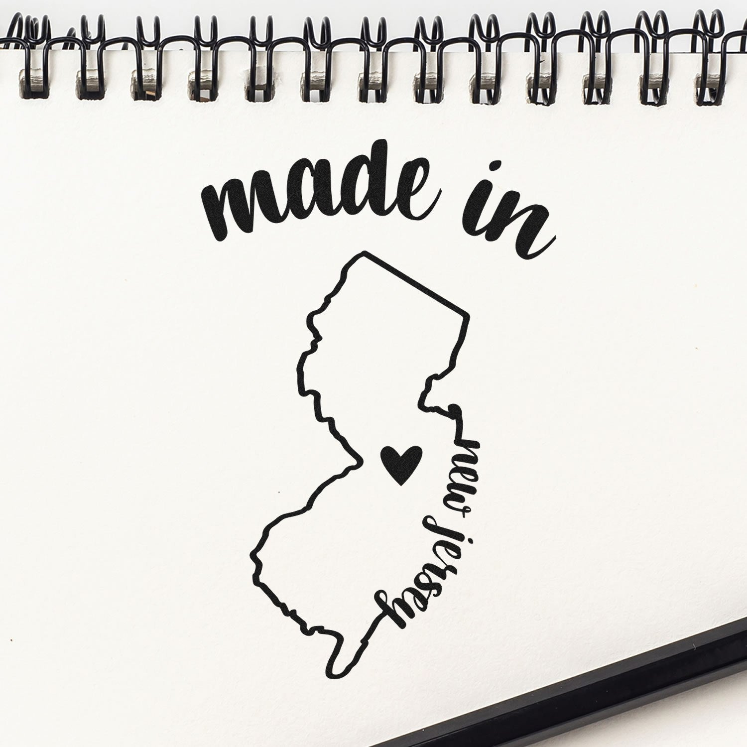 Made in New Jersey Stamp Pre-Inked featuring a black outline of New Jersey with a heart, on a spiral notebook. Perfect for adding a local touch to crafts and documents.