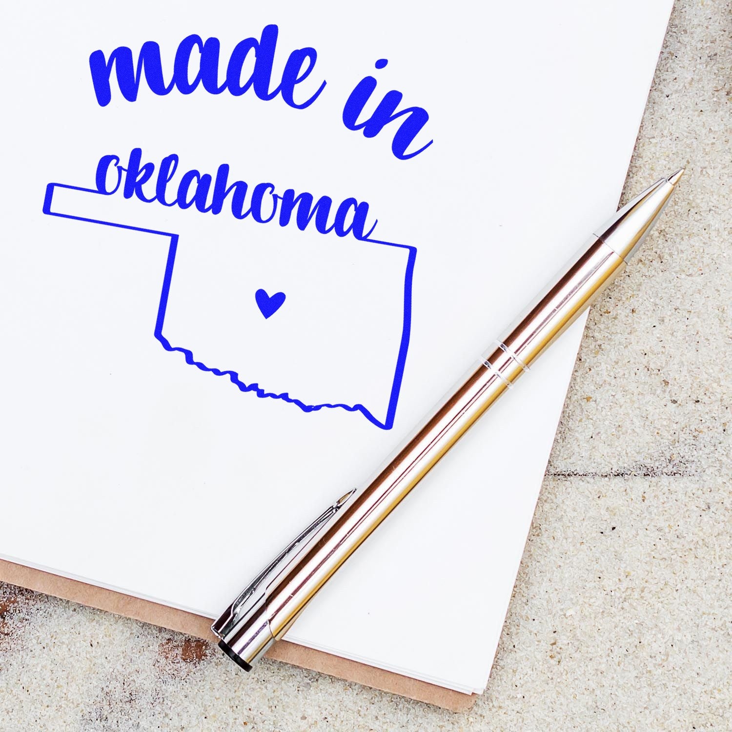 Made with Love in Oklahoma Rubber Stamp featuring a blue outline of Oklahoma with a heart, placed on a white paper next to a gold pen.