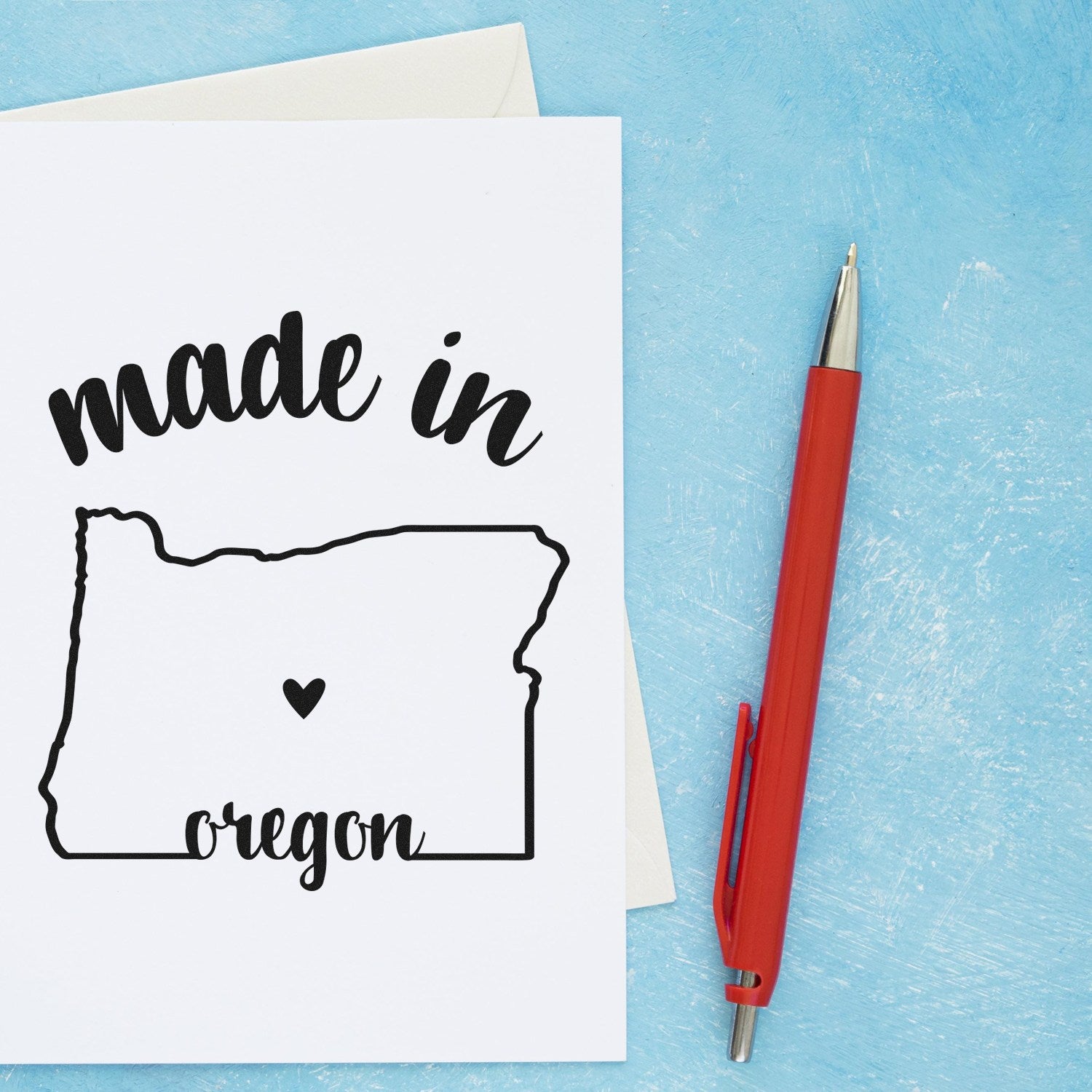 Self-Inking Handmade with Love in Oregon Stamp on a card with a red pen, featuring a heart inside the Oregon state outline and 'made in oregon' text on a blue background.