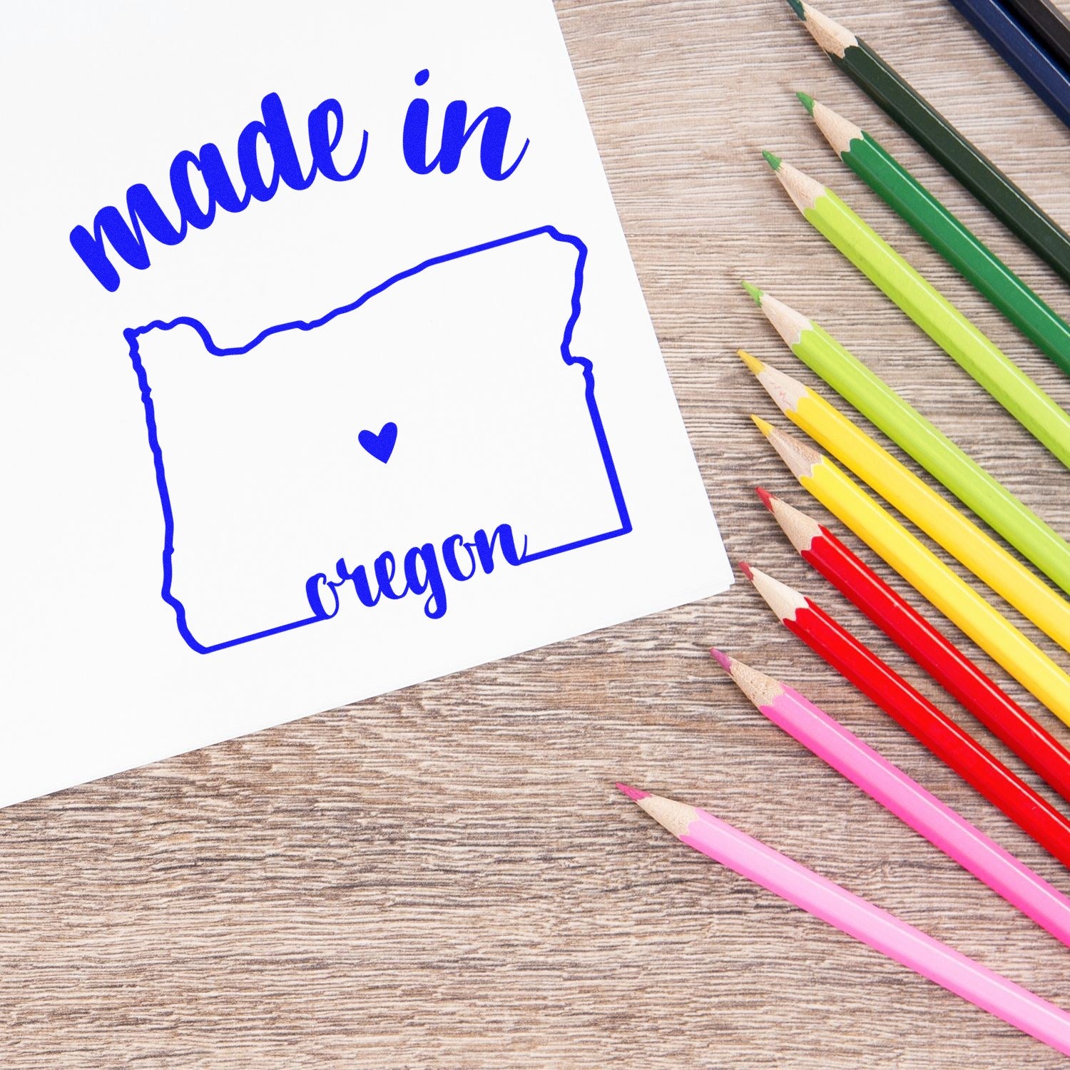 Self-Inking Handmade with Love in Oregon Stamp featuring a blue outline of Oregon with a heart, surrounded by colorful pencils on a wooden surface.