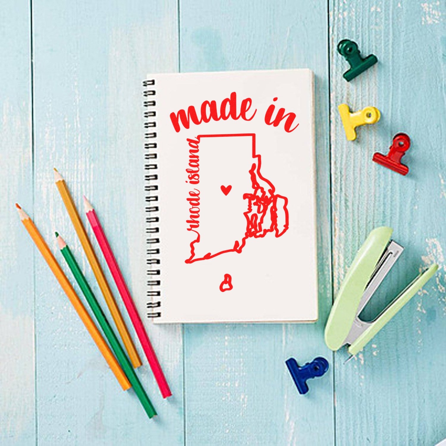 Made with Love in Rhode Island Rubber Stamp on a notebook, featuring a red outline of Rhode Island with hearts, surrounded by colorful pencils and clips on a blue wooden surface.