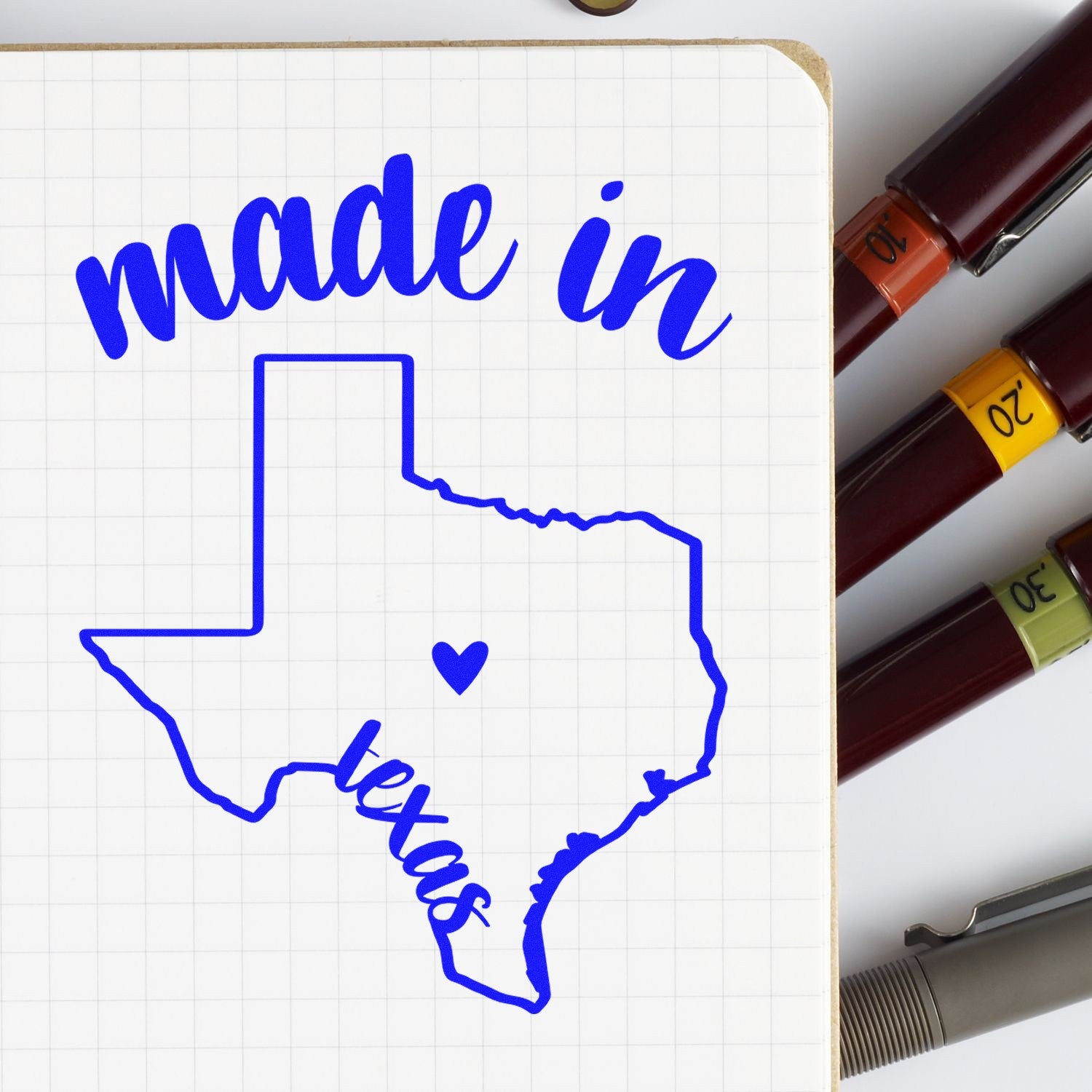 Self-Inking Handmade with Love in Texas Stamp featuring a blue Texas outline with made in Texas text and heart, shown on a notebook with pens. Perfect for crafts and gifts.