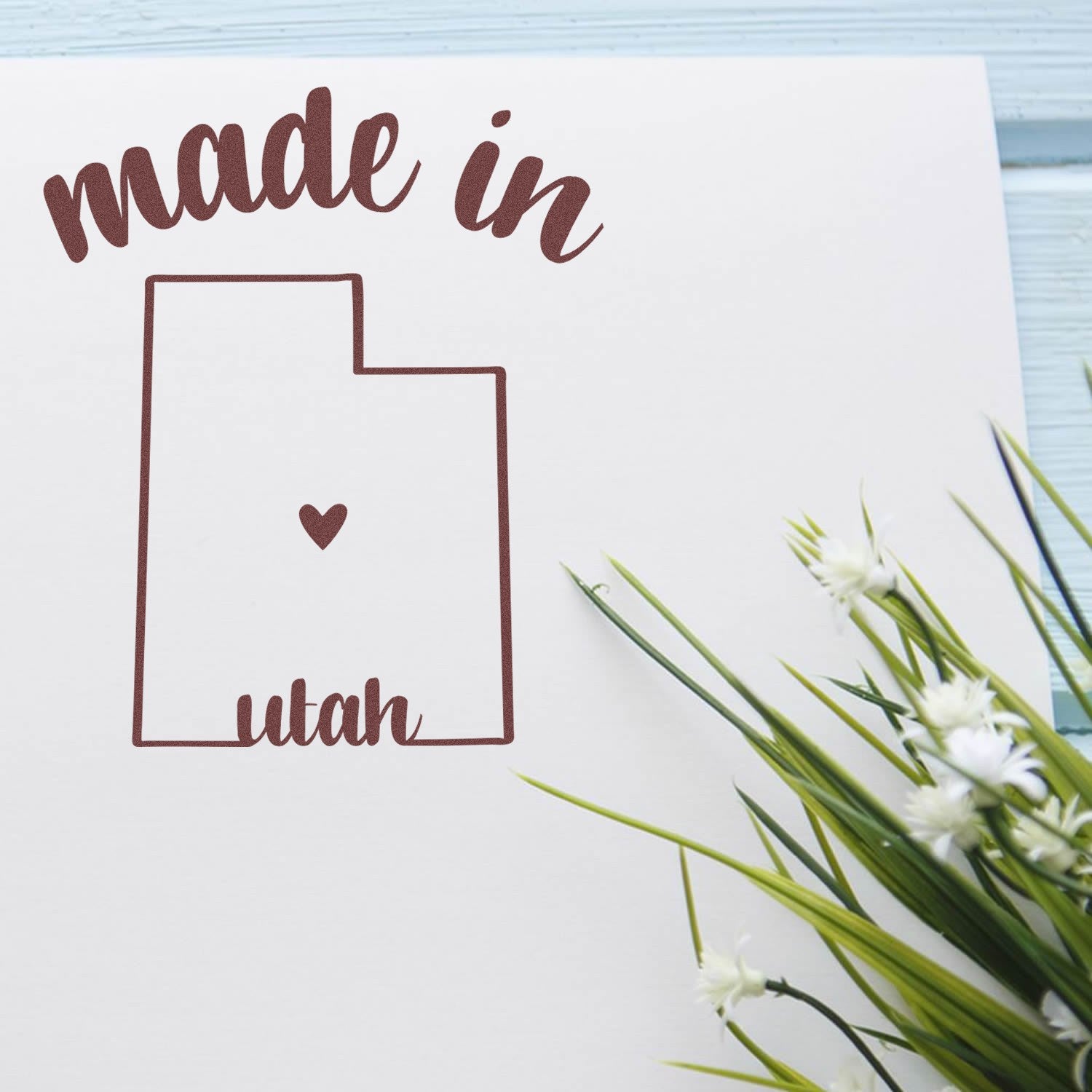 Made in Utah Stamp Pre-Inked featuring a heart inside the Utah state outline, with 'made in' text above and 'Utah' below, on white paper next to green plant.