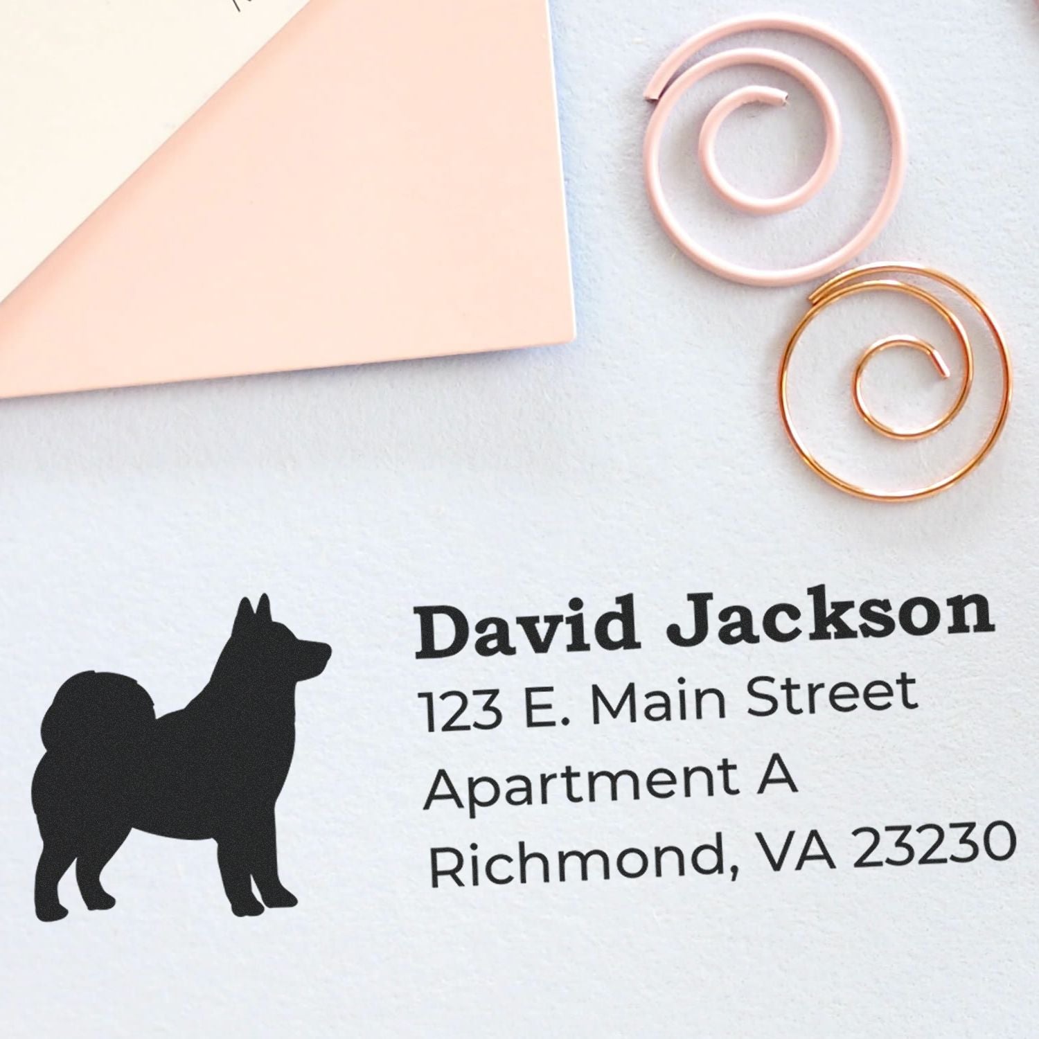 Alaskan Malamute Silhouette Address Rubber Stamp on paper with a silhouette of a dog, personalized with name and address. Nearby are pink and gold spiral paper clips and an envelope corner.