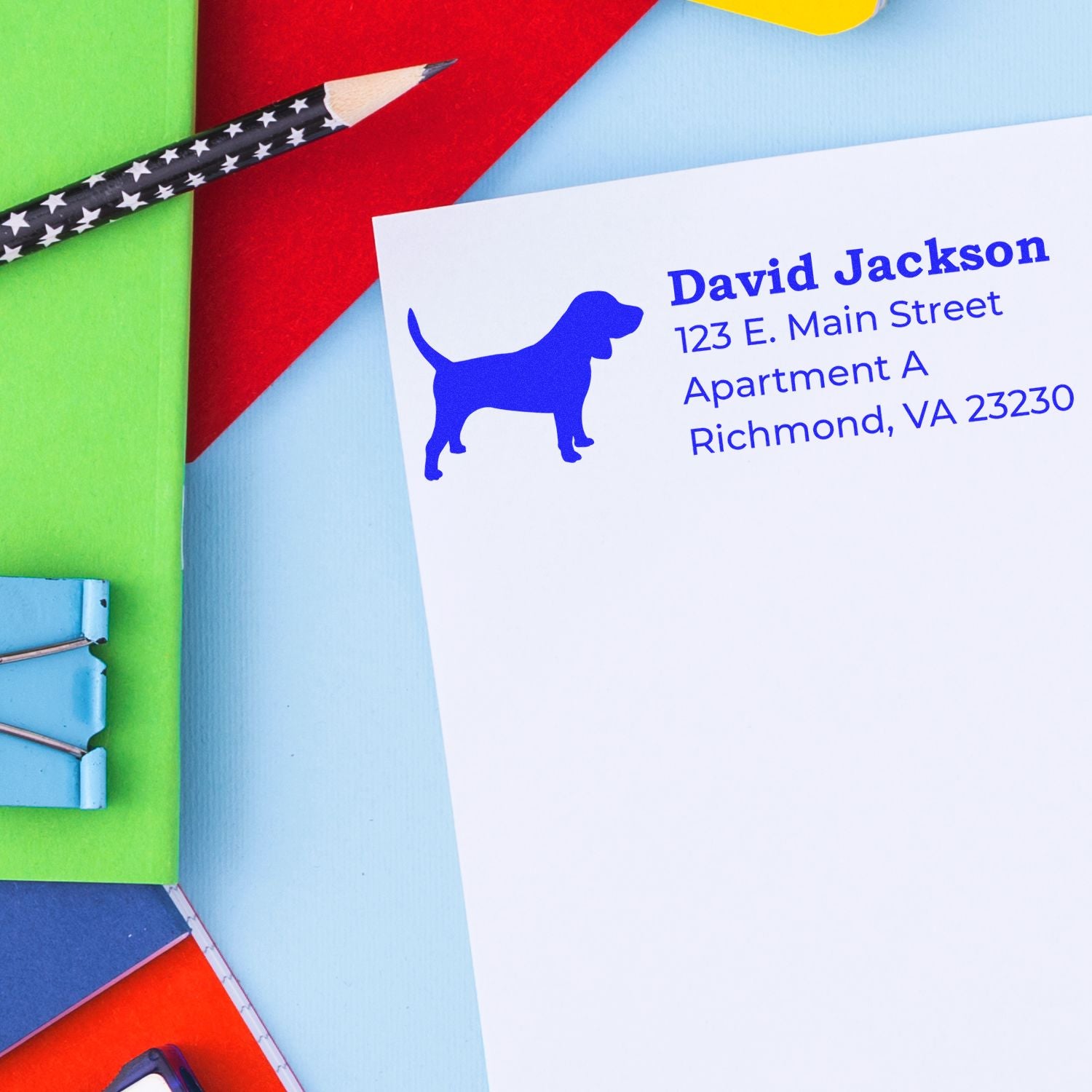 A colorful desk with a Slim Pre-Inked Beagle Address Stamp imprint on a white envelope, featuring a blue beagle silhouette and address details.