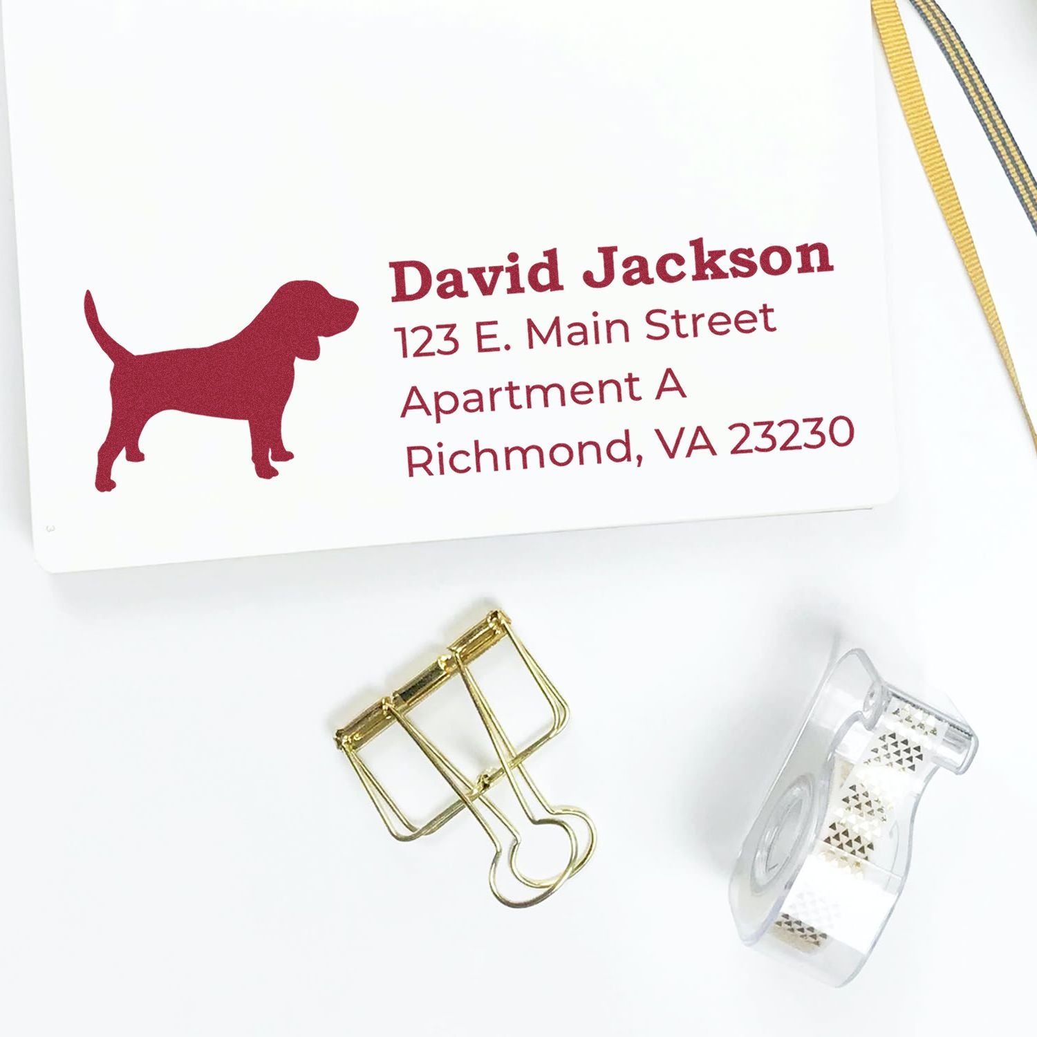 Slim Pre-Inked Beagle Address Stamp on white paper with a red beagle silhouette and sample address. Nearby are a gold binder clip and decorative tape, creating a stylish and organized workspace.