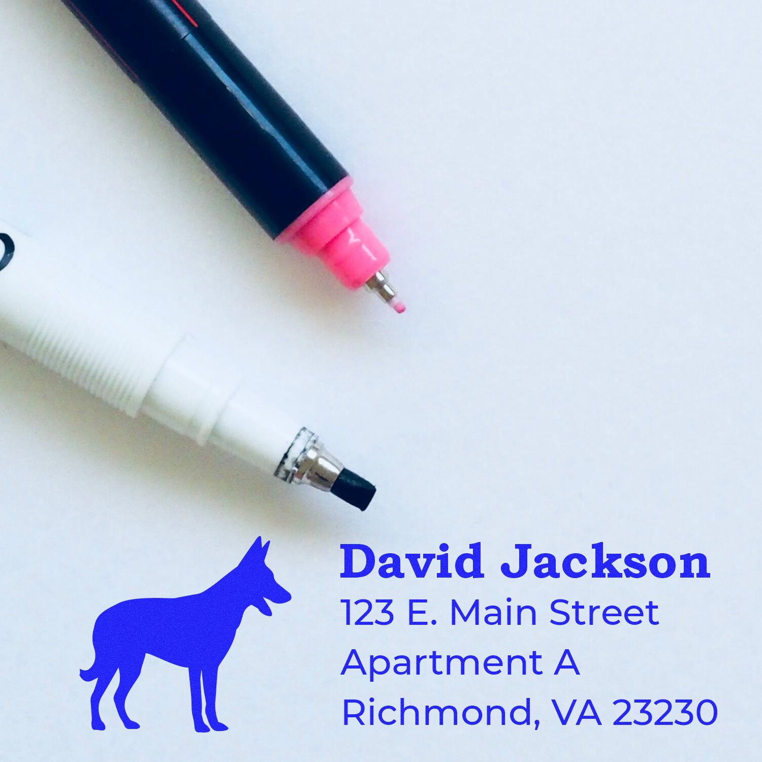 Slim Pre-Inked Belgian Malinois Address Stamp in use, featuring a blue dog silhouette and address details on white paper, with two pens nearby. Perfect for personalized mailings.