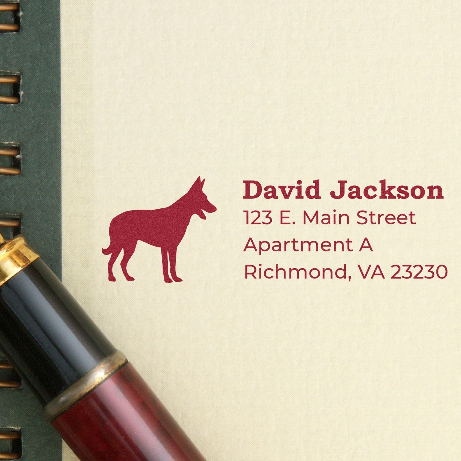 Self-Inking Belgian Malinois Dog Address Stamp on paper with a red pen nearby. The stamp features a silhouette of a Belgian Malinois and sample address text in red ink.