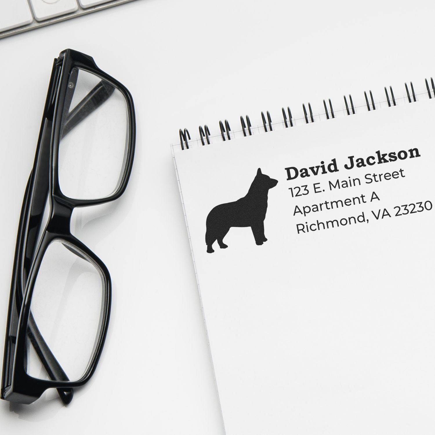 Notebook with Blue Heeler Silhouette Address Rubber Stamp imprint, showing a silhouette of a dog next to an address. Glasses and keyboard nearby, creating a professional desk setting.